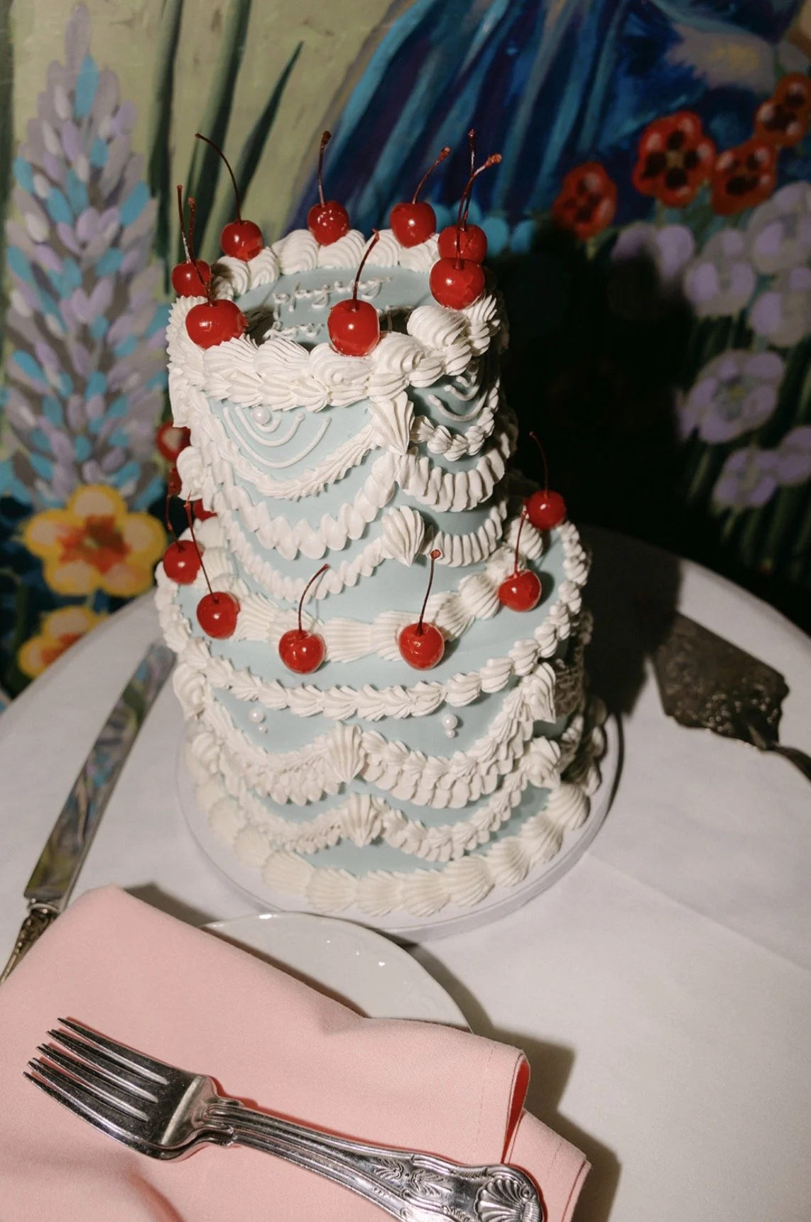 Tiered birthday cake decorated with white icing, cherry toppings, and blue and white swirling icing patterns, placed on a table with a pink napkin and fork, with colorful floral and landscape background.