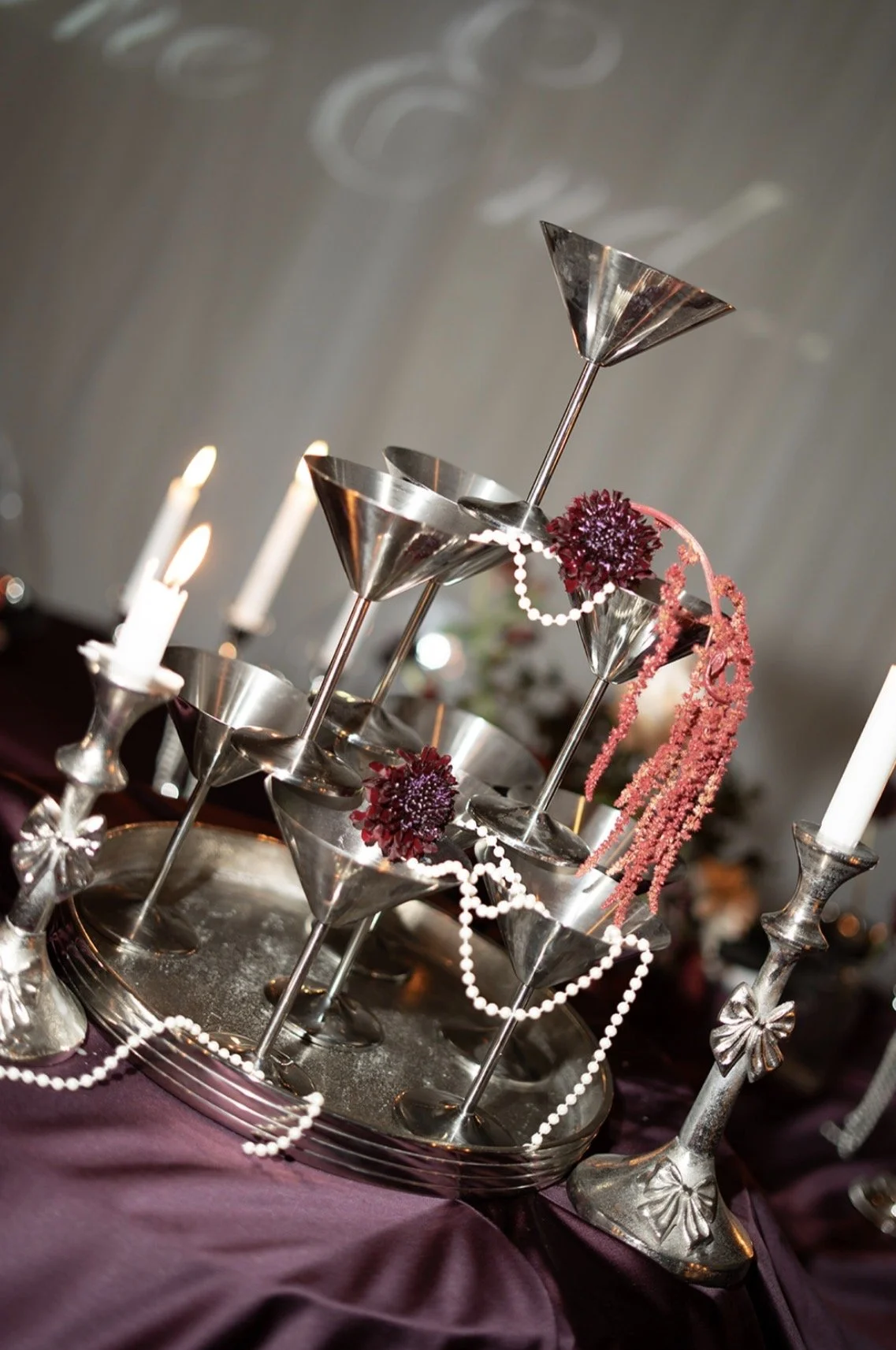 A decorative silver and metal centerpiece with martini glasses, candles, pink flowers, and strings of pearls on a purple tablecloth