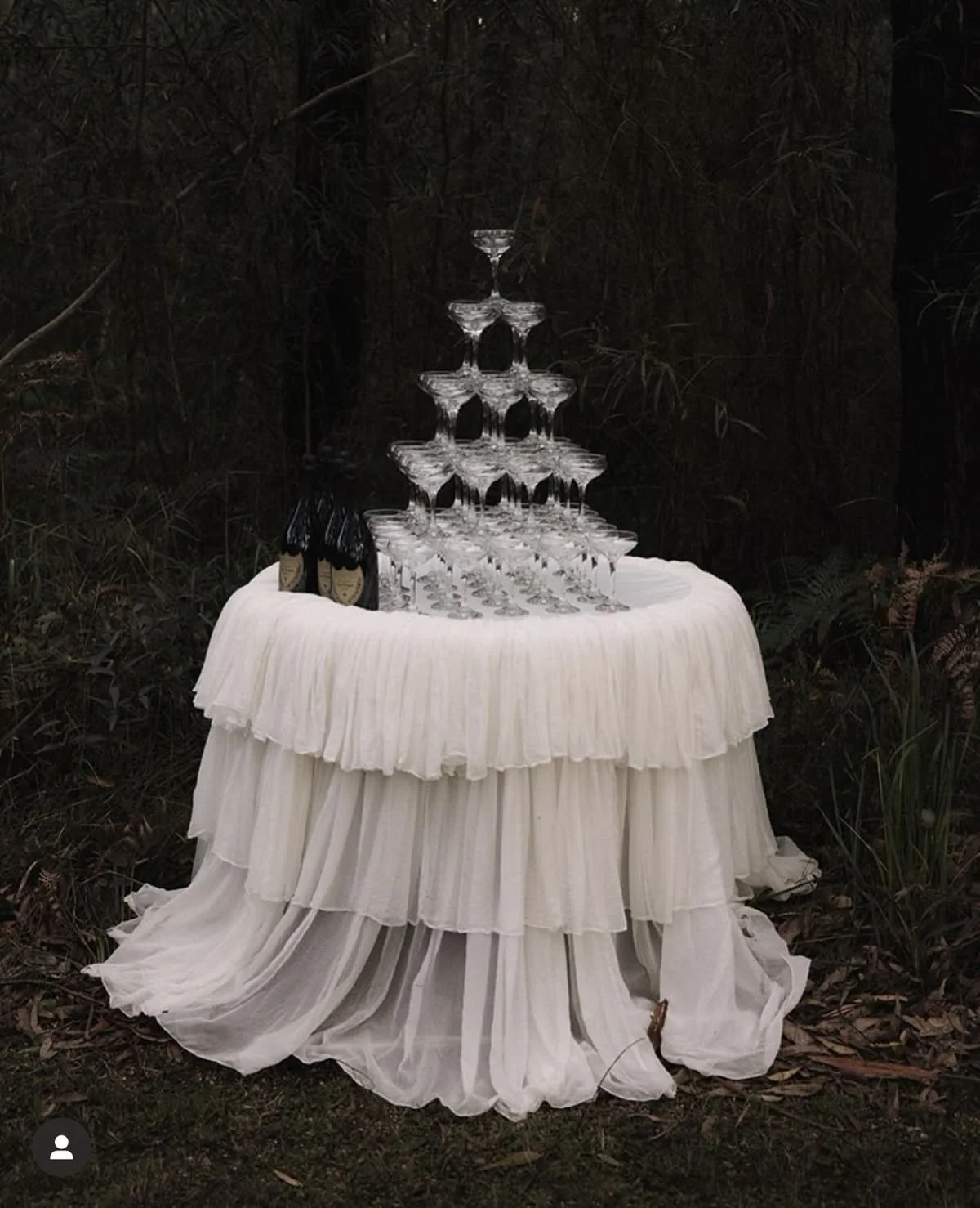 A tiered champagne glass tower on a white ruffled tablecloth with a forest background.