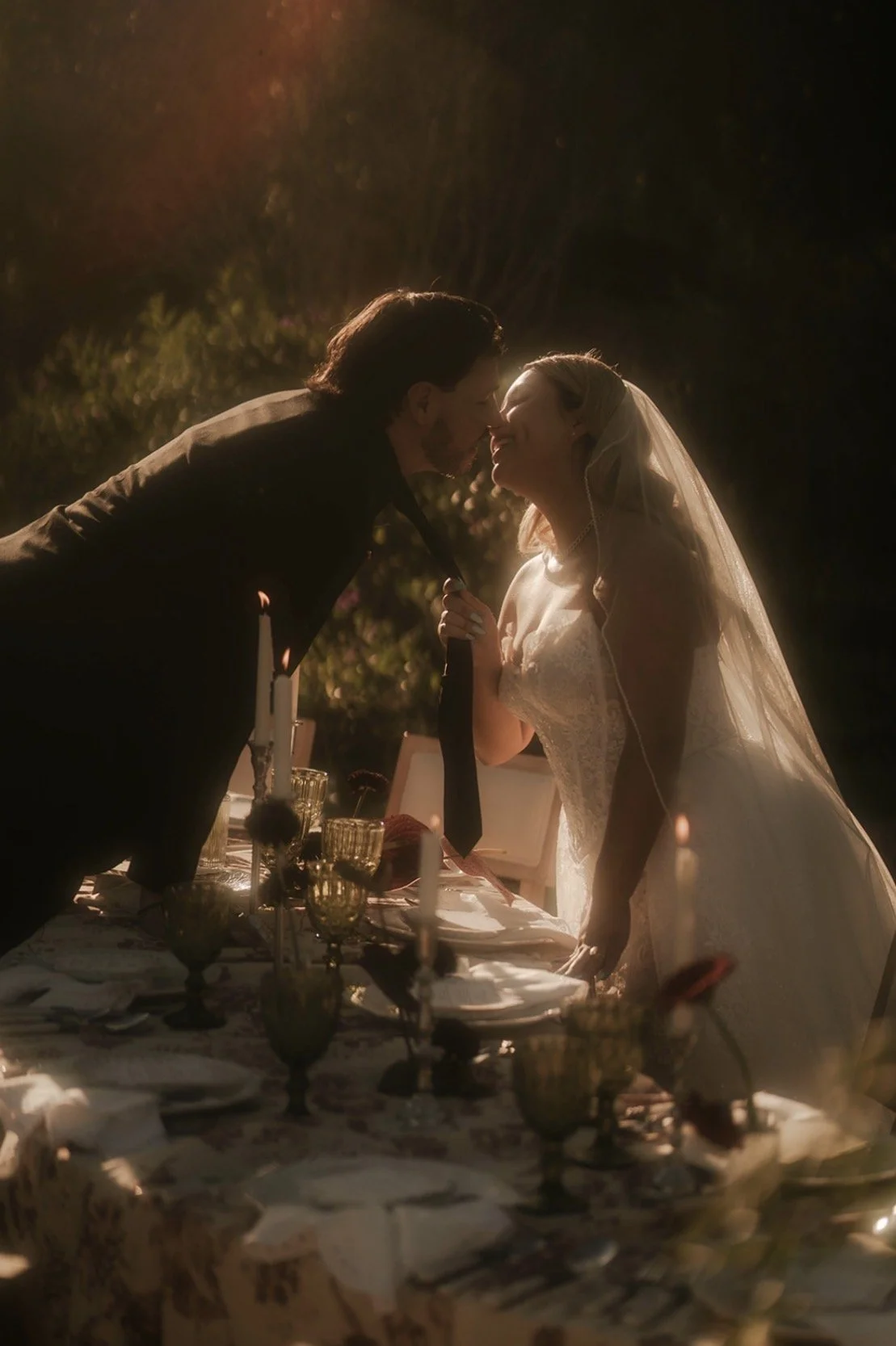 A bride and groom leaning close to each other during their wedding dinner, touching noses, illuminated warm light at night.