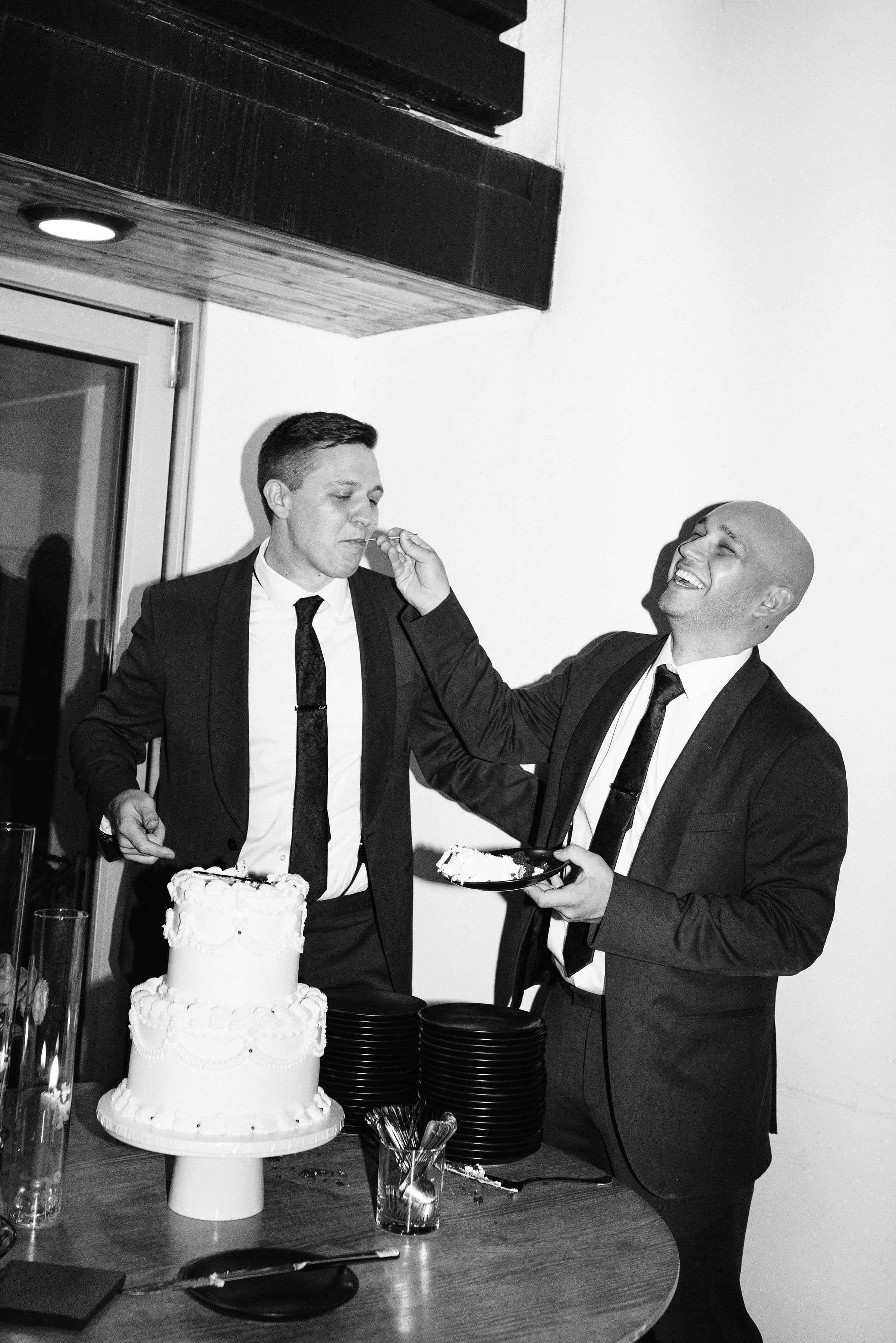 Two men in suits at a celebration, with a cake on the table, one feeding the other a piece of cake while smiling and laughing, in black and white.