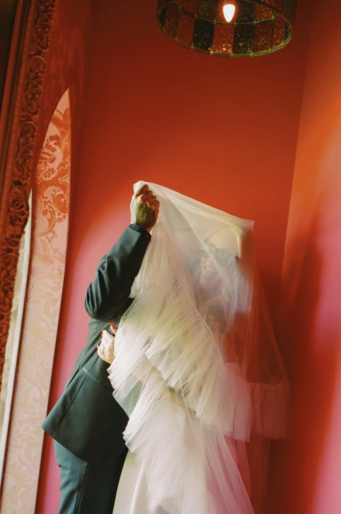 A groom in a dark suit partially hidden behind a bride in a white wedding dress with a veil, in front of a red wall with ornate decor and a hanging light.