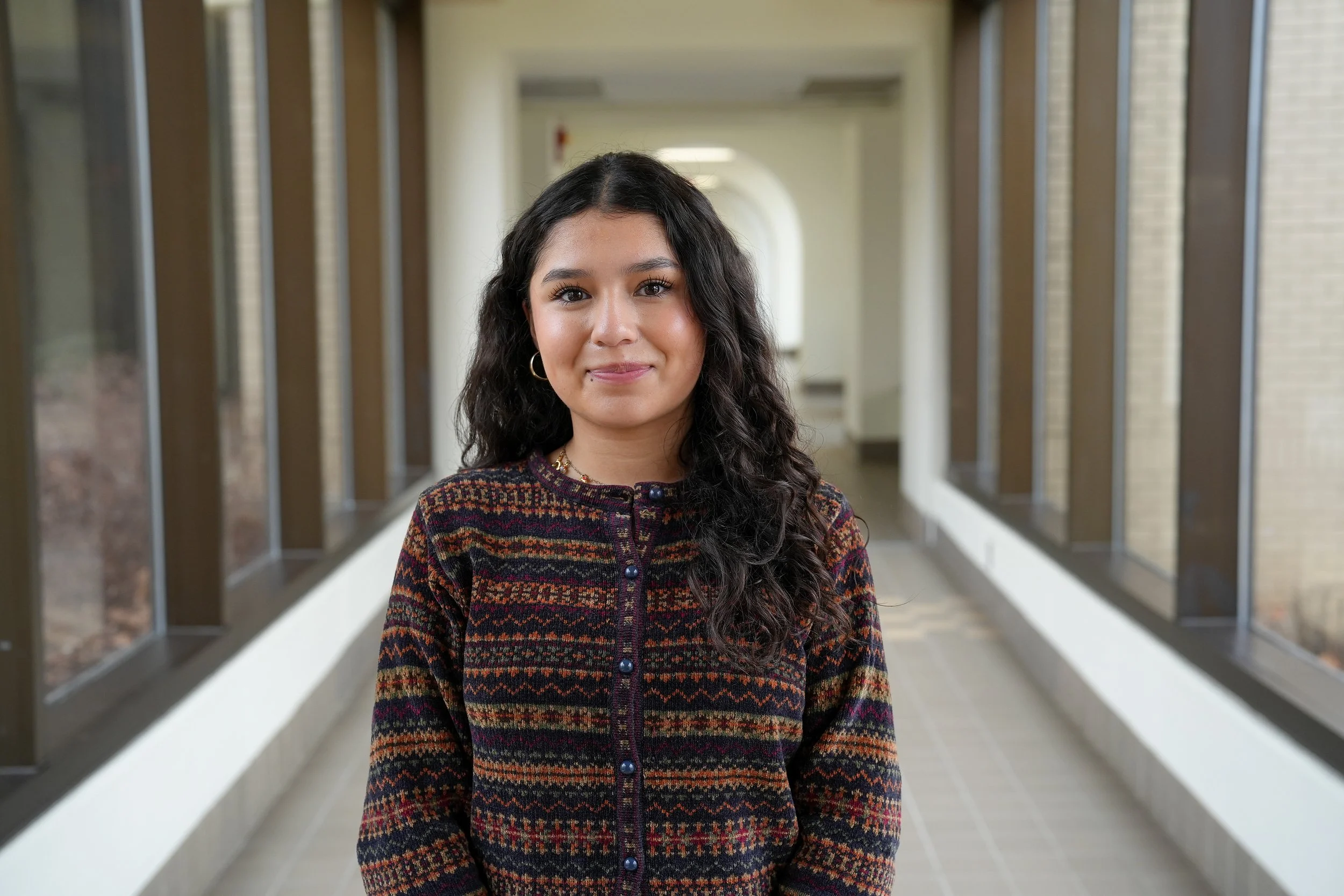Young woman with dark curly hair smiling, standing in a hallway with large windows and brick exterior walls.