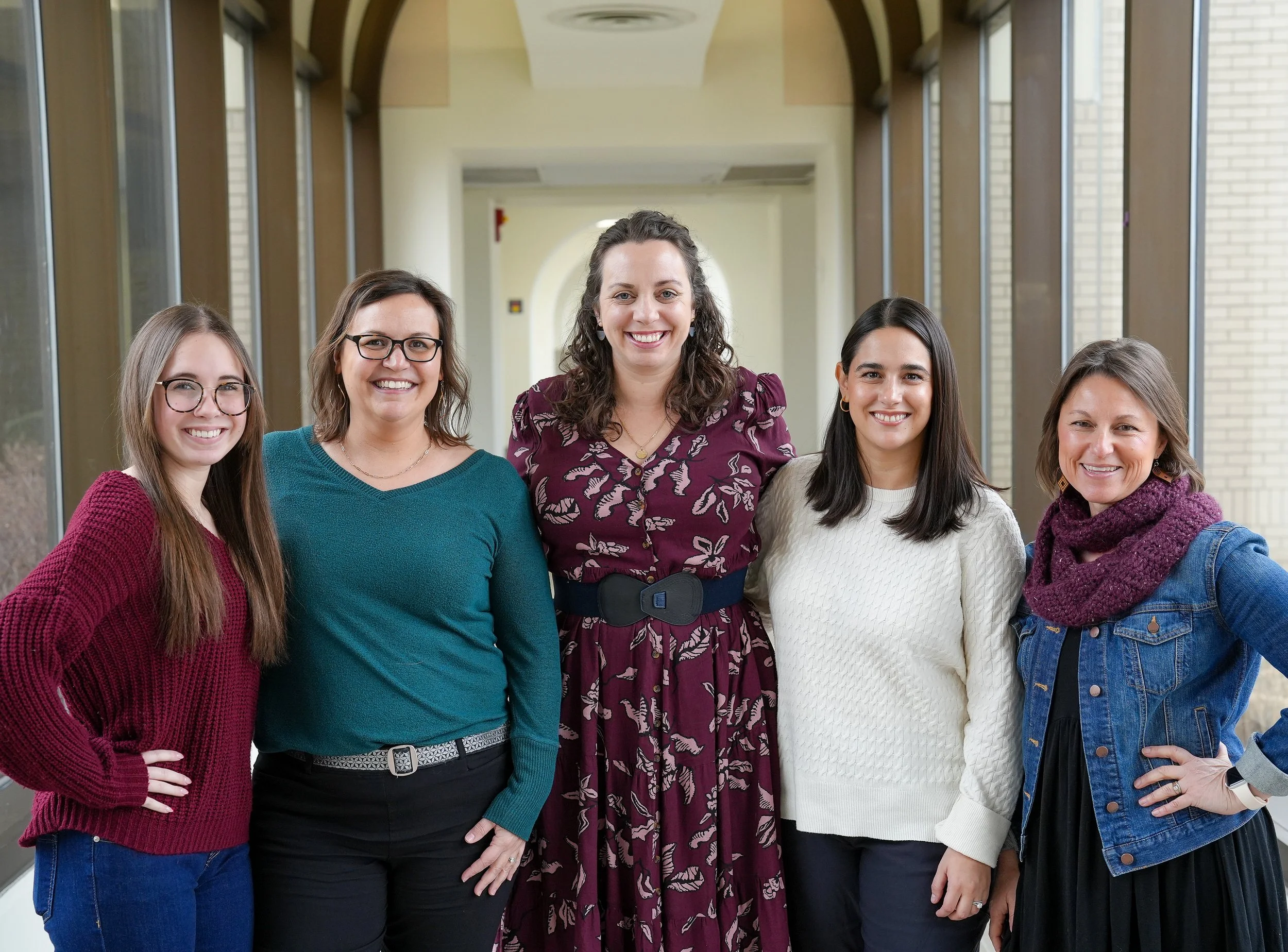 Group of five women standing together in a brightly lit indoor hallway, smiling at the camera.