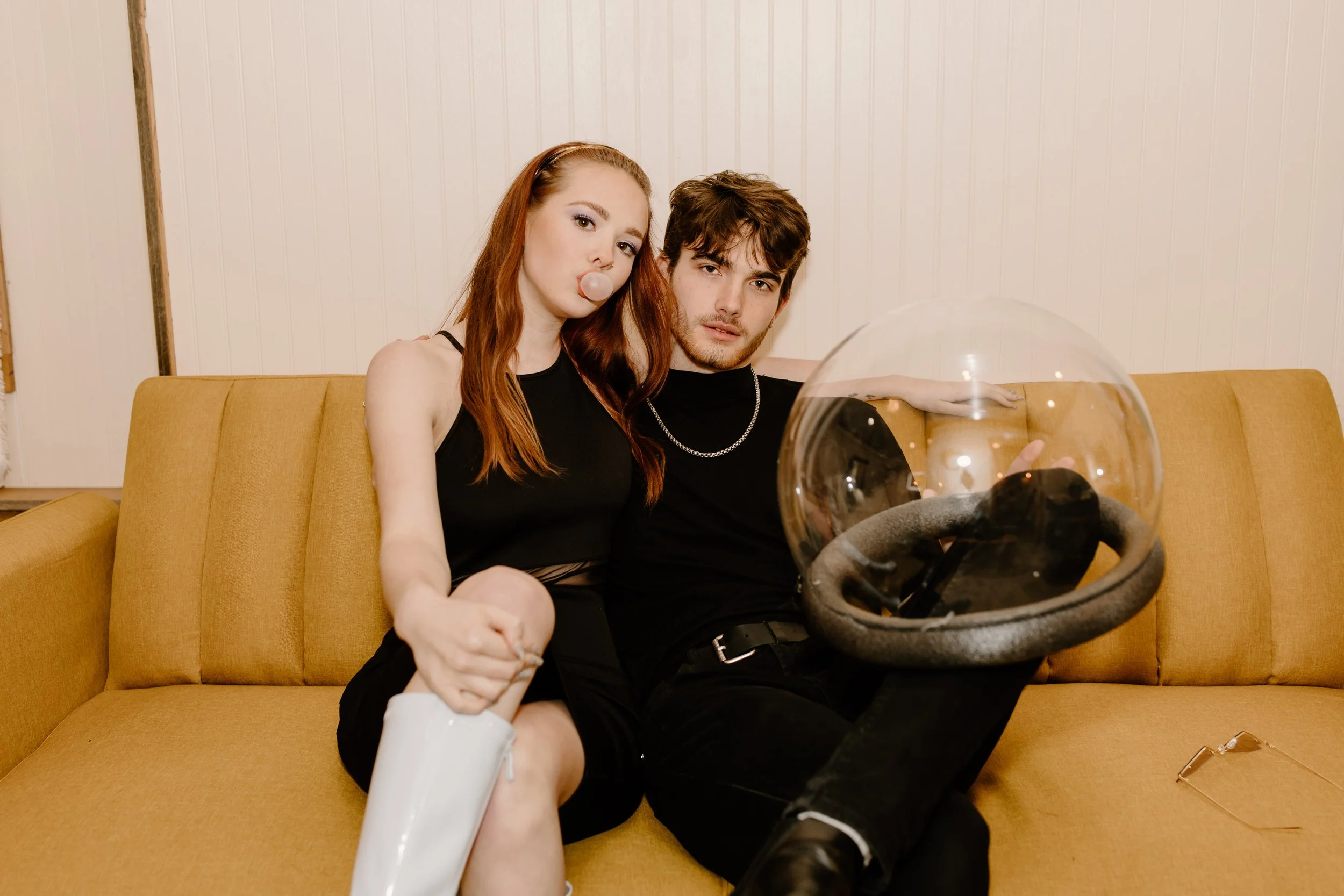 A young woman with red hair and a young man with brown hair sitting on a yellow sofa in a room with white paneled walls. The woman is blowing a bubble gum bubble, wearing a black sleeveless top, and holding a white bottle. The man is wearing a black shirt, a silver chain, and black pants. There is a large transparent glass orb near him, and a pair of glasses on the sofa near the man's foot.