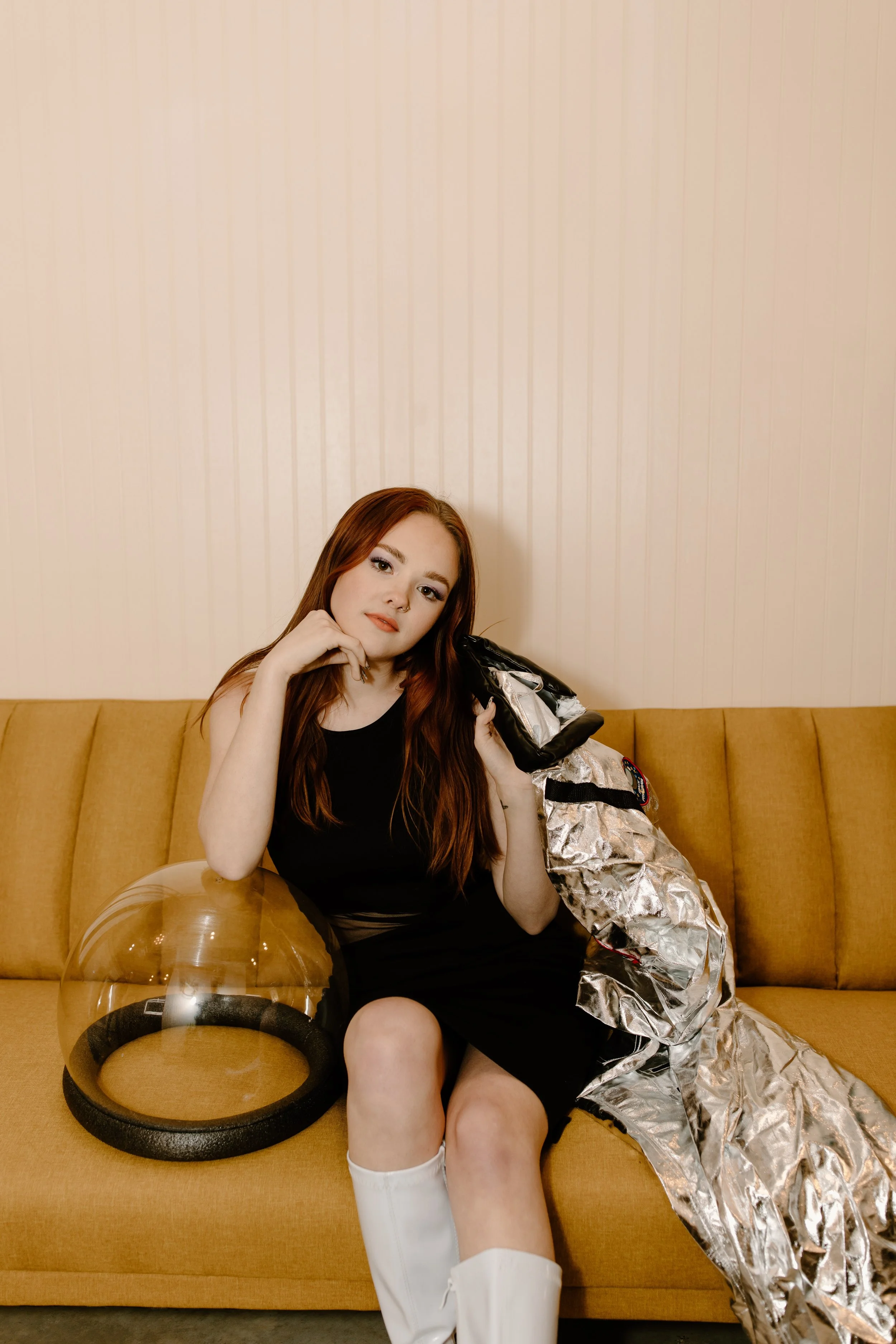 A woman with long red hair sitting on a yellow couch, wearing a black dress and white knee-high boots, holding a silver space suit and helmet.