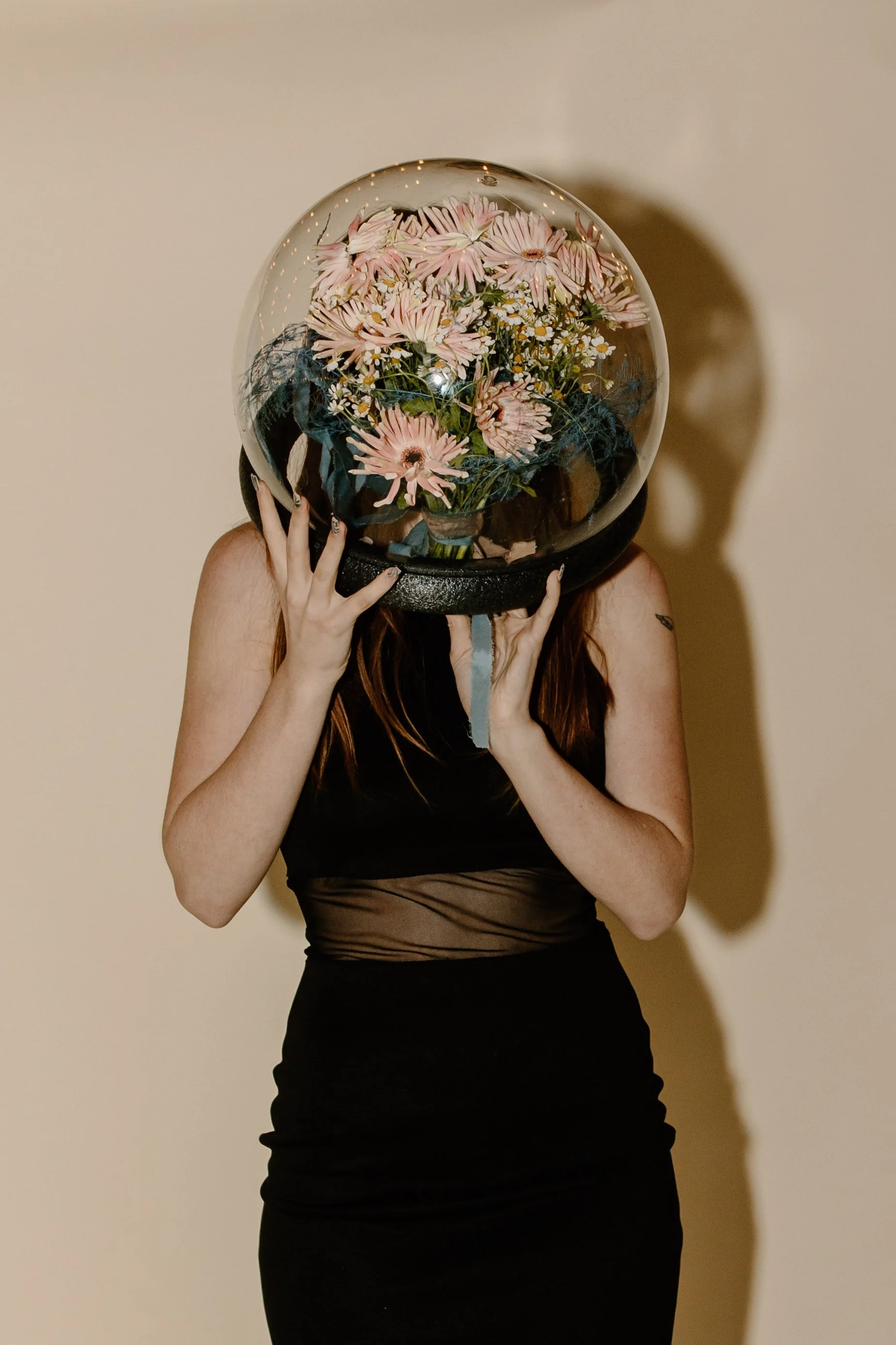 Person holding a glass globe filled with pink and white flowers in front of their face, standing against a plain wall.