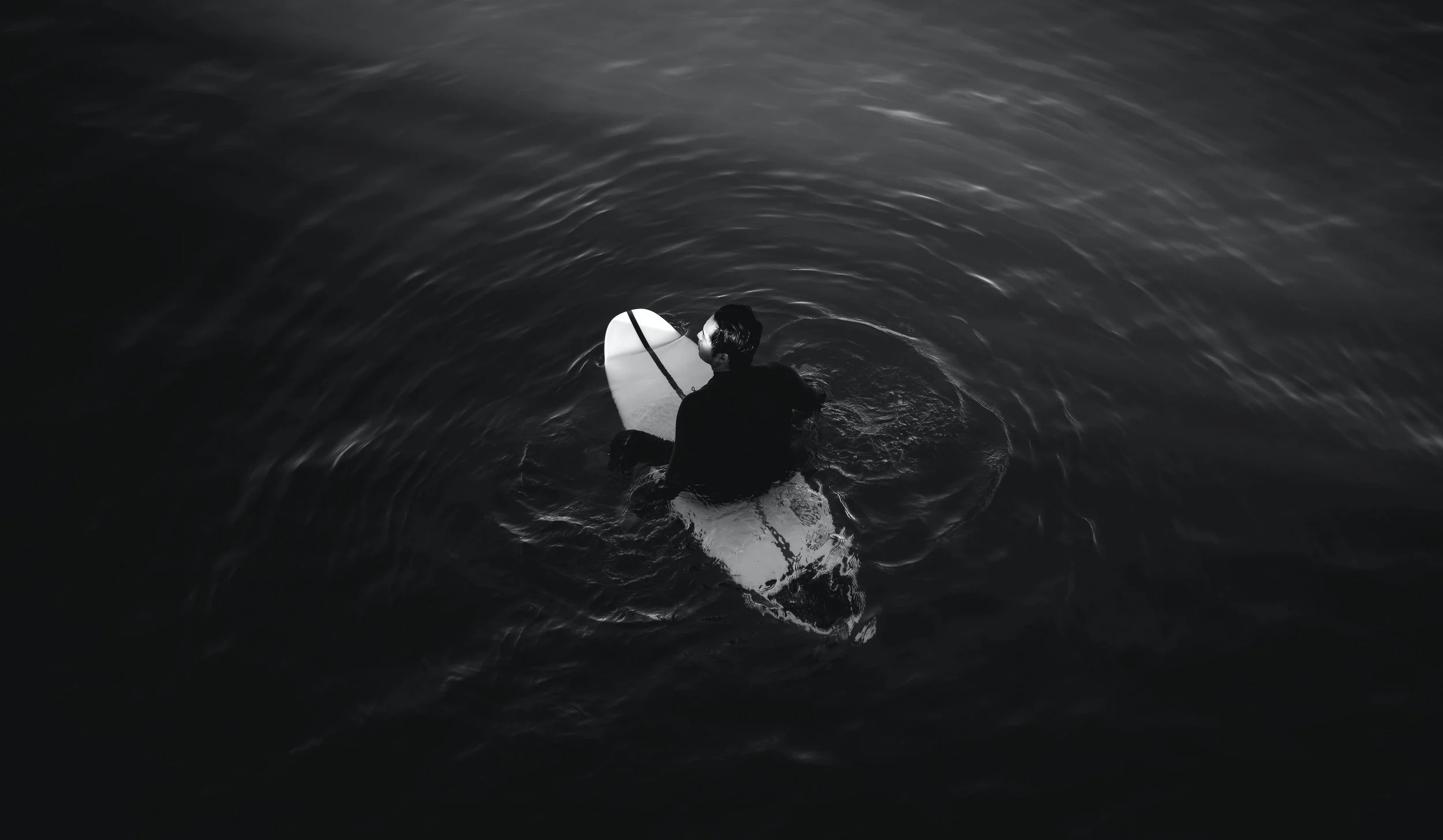 A man in a dark wetsuit paddling a surfboard in dark water, viewed from above.
