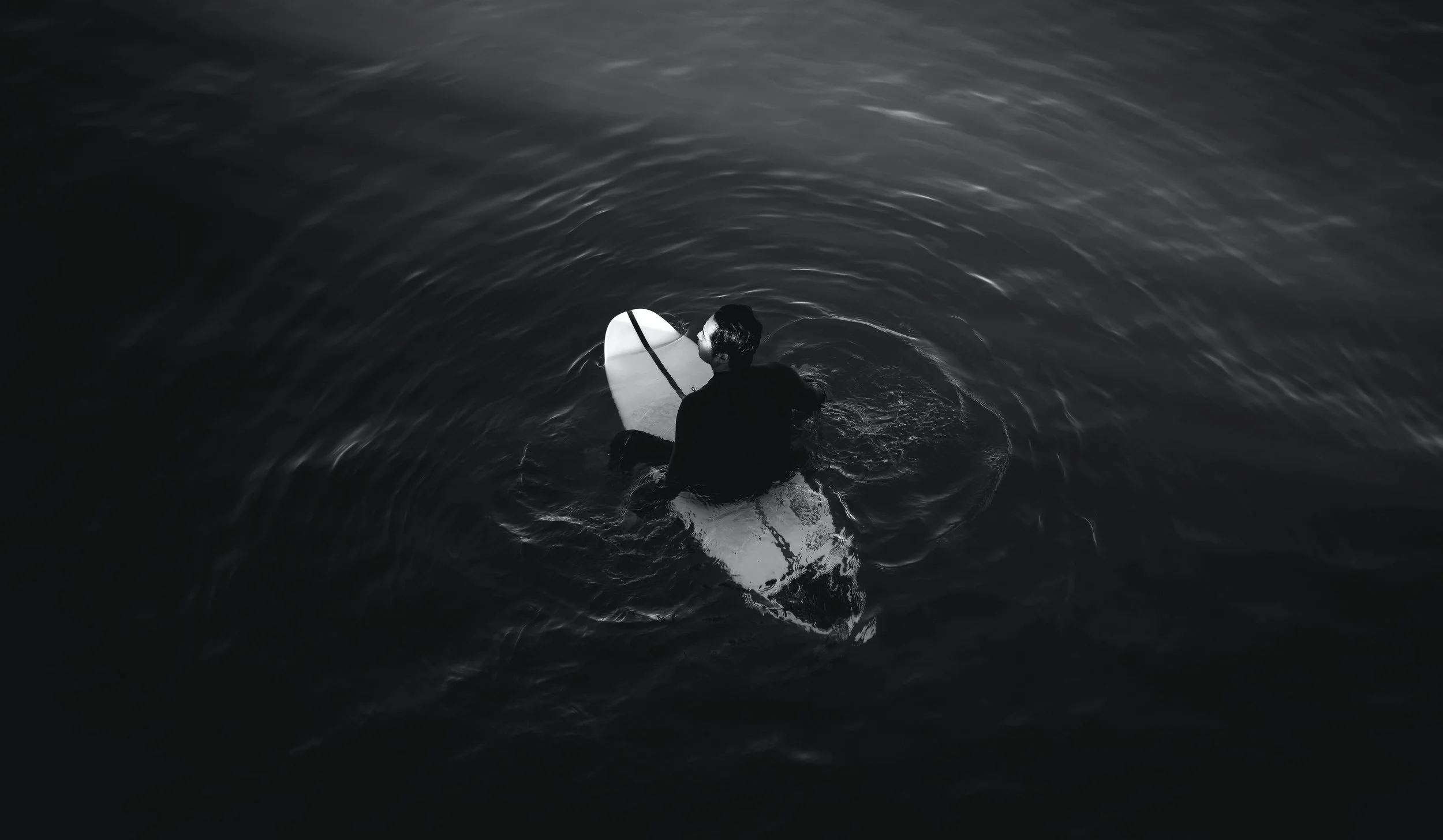 A person with dark hair, dressed in black, sitting on a surfboard in a dark body of water, viewed from above.