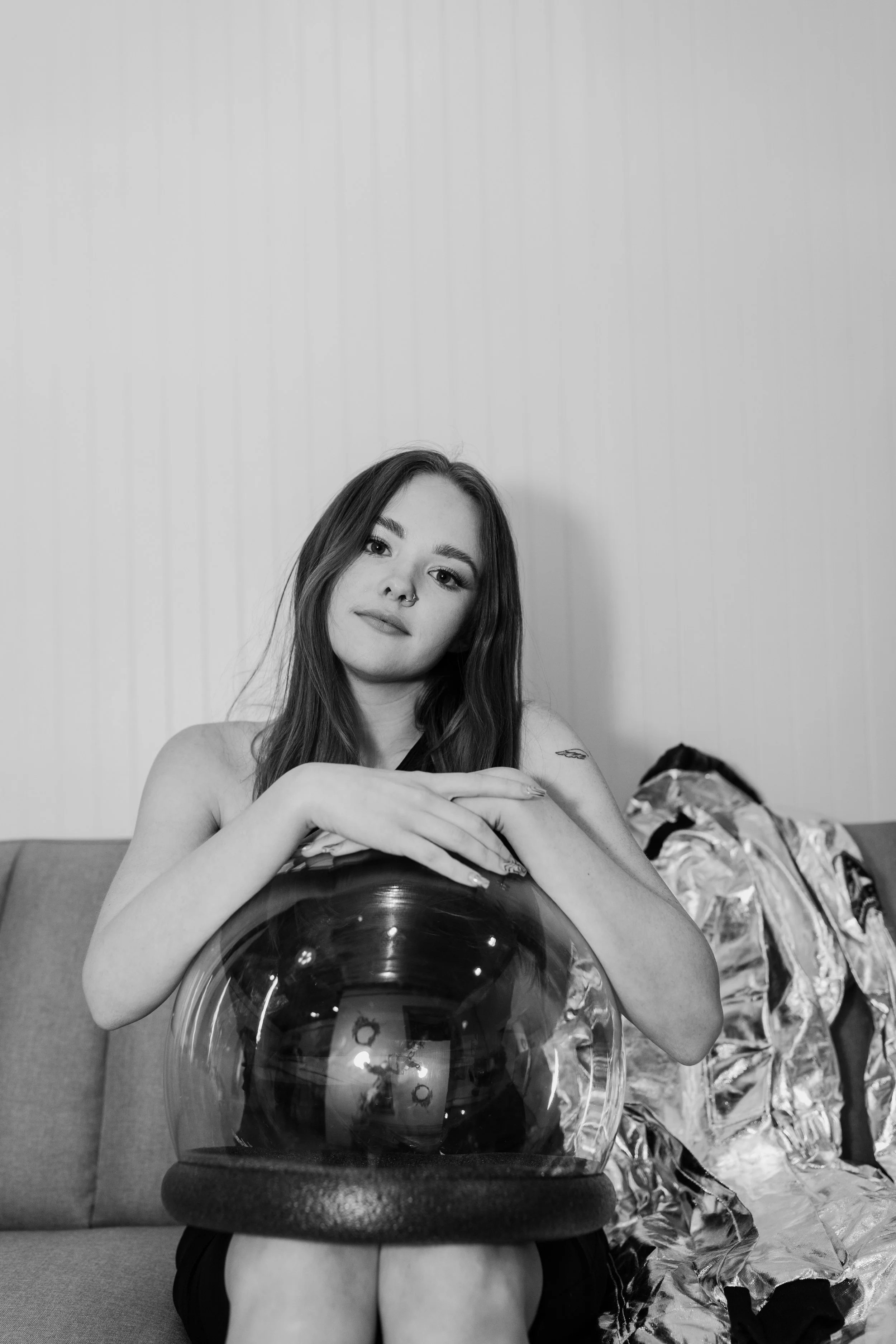 A young woman with long hair sitting on a couch, holding a glass sphere with reflections inside, and looking at the camera with a neutral expression. There are clothes in a bag next to her on the couch.