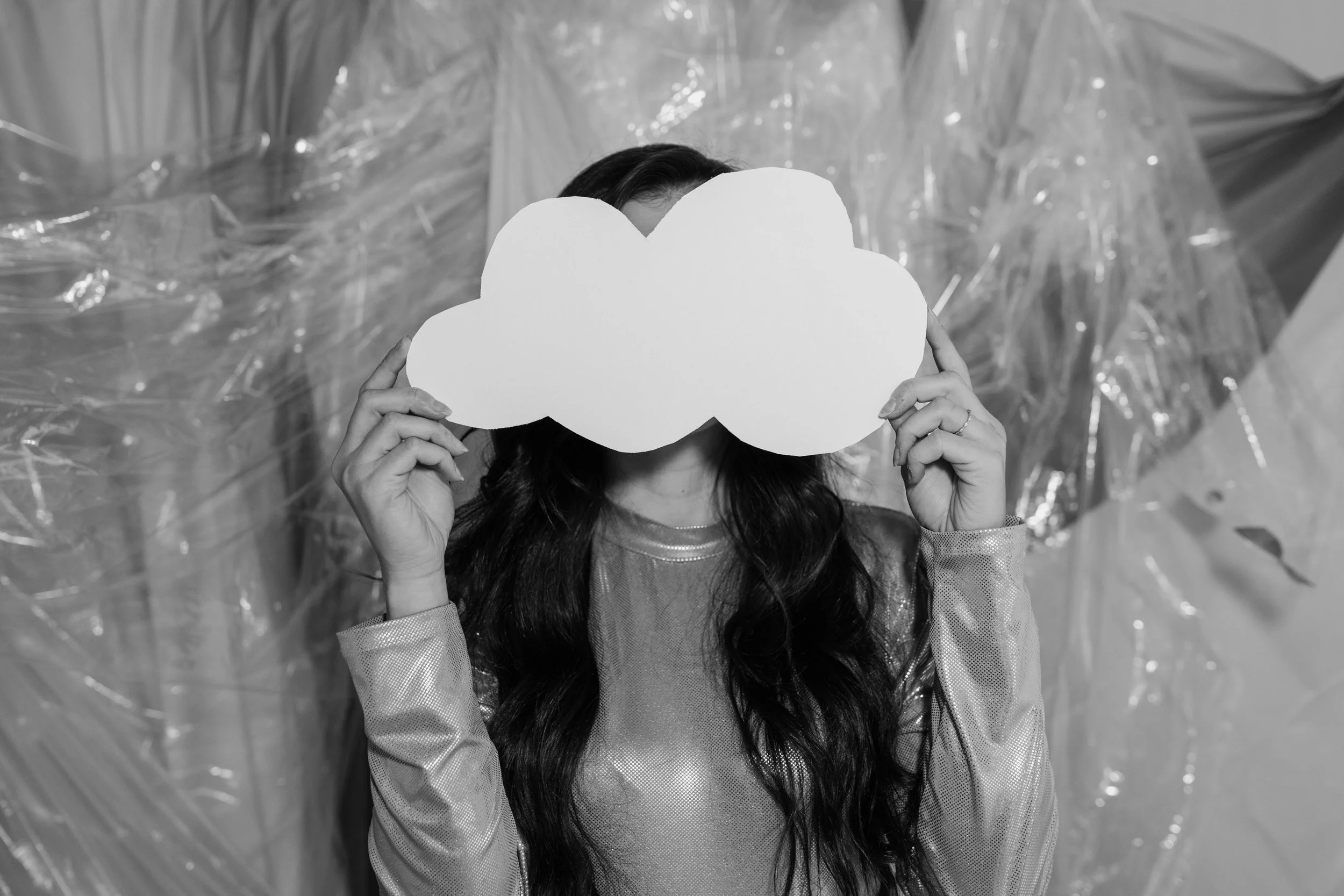 A woman with long dark hair holding a cloud-shaped cutout in front of her face, standing in front of a textured background.