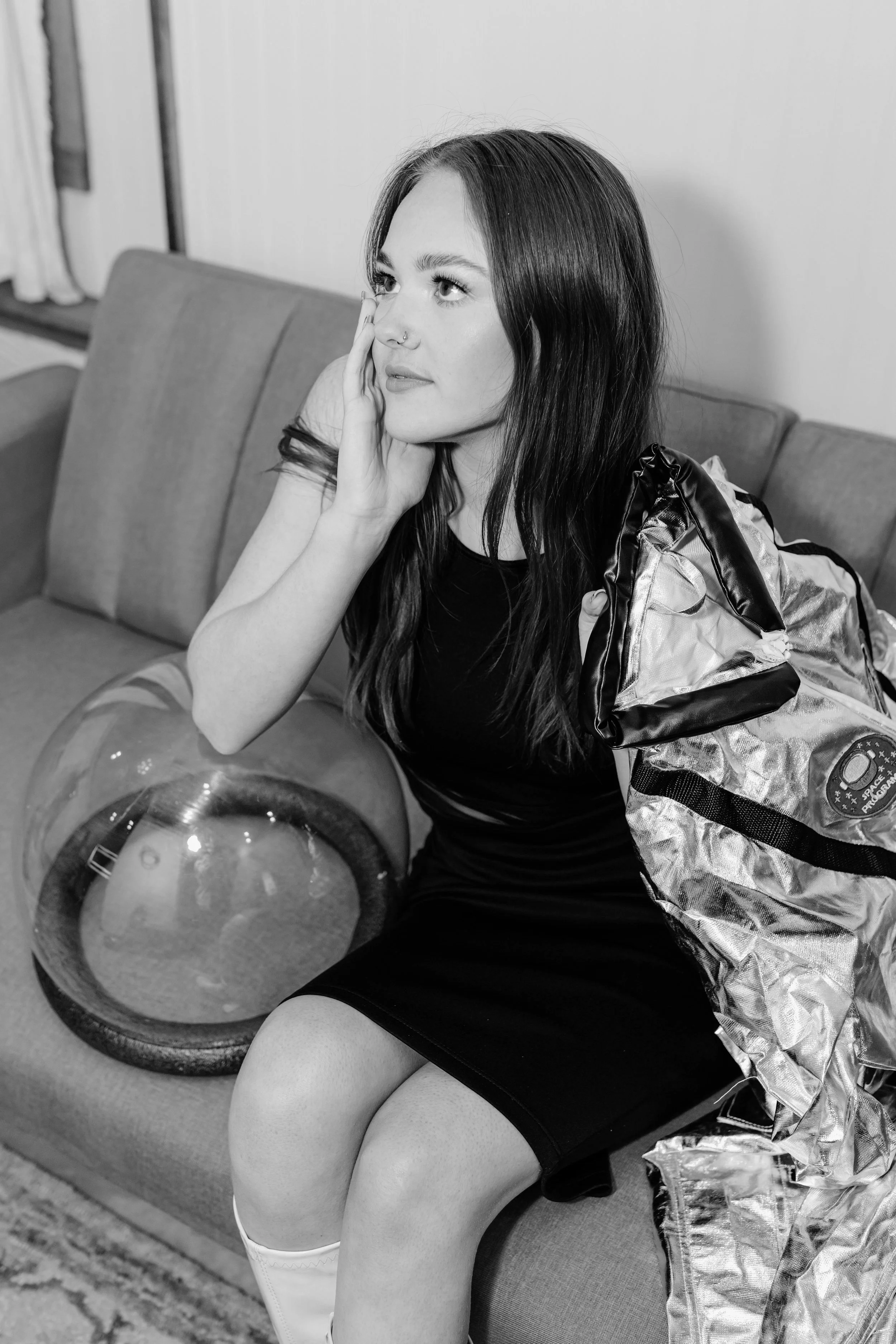 A woman with long dark hair sitting on a couch, resting her face on her hand, with a reflective space blanket draped over her shoulder and a large glass container with water next to her.