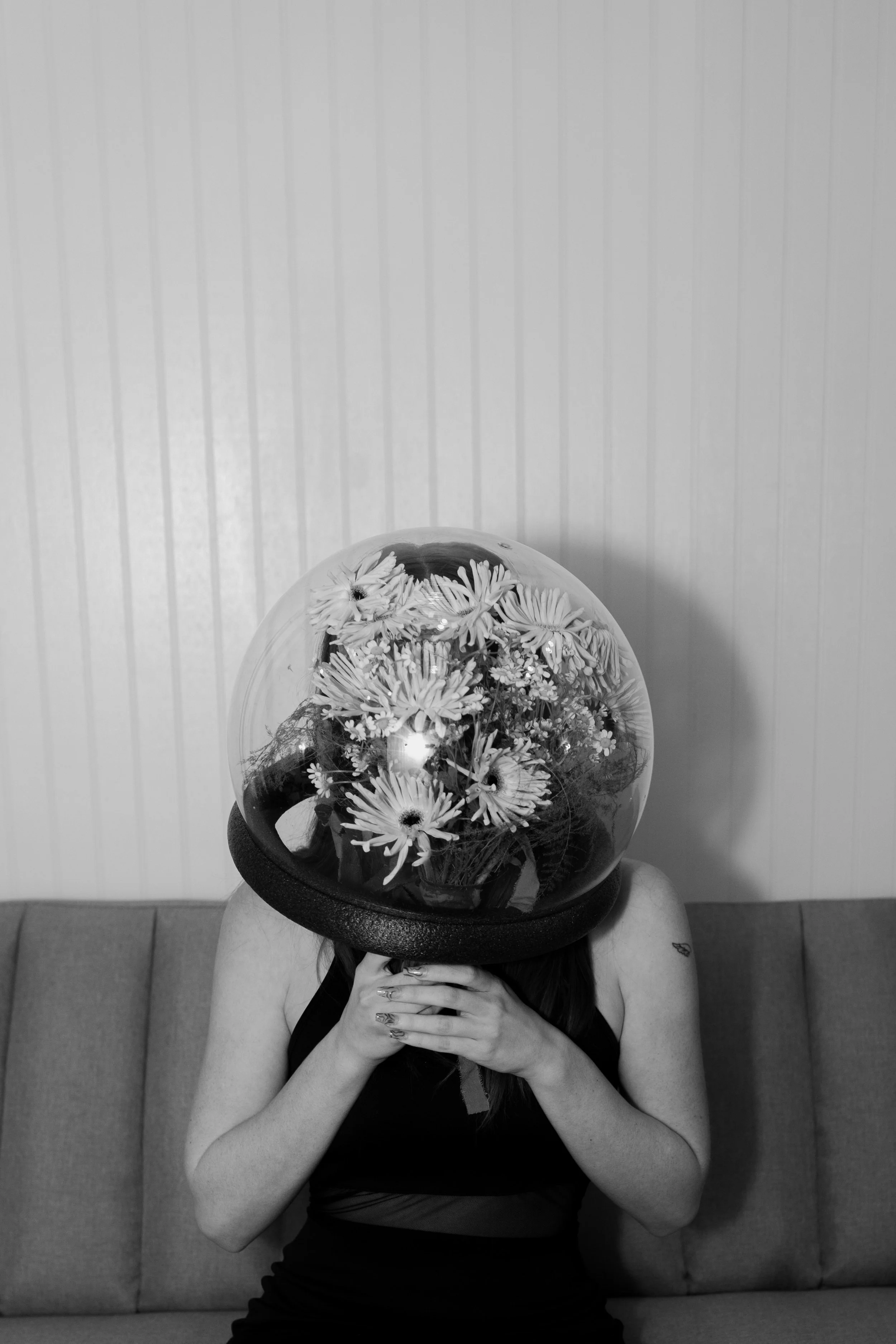 A woman with a balloon filled with flowers covering her face, sitting on a couch against a wall with vertical striped wallpaper.