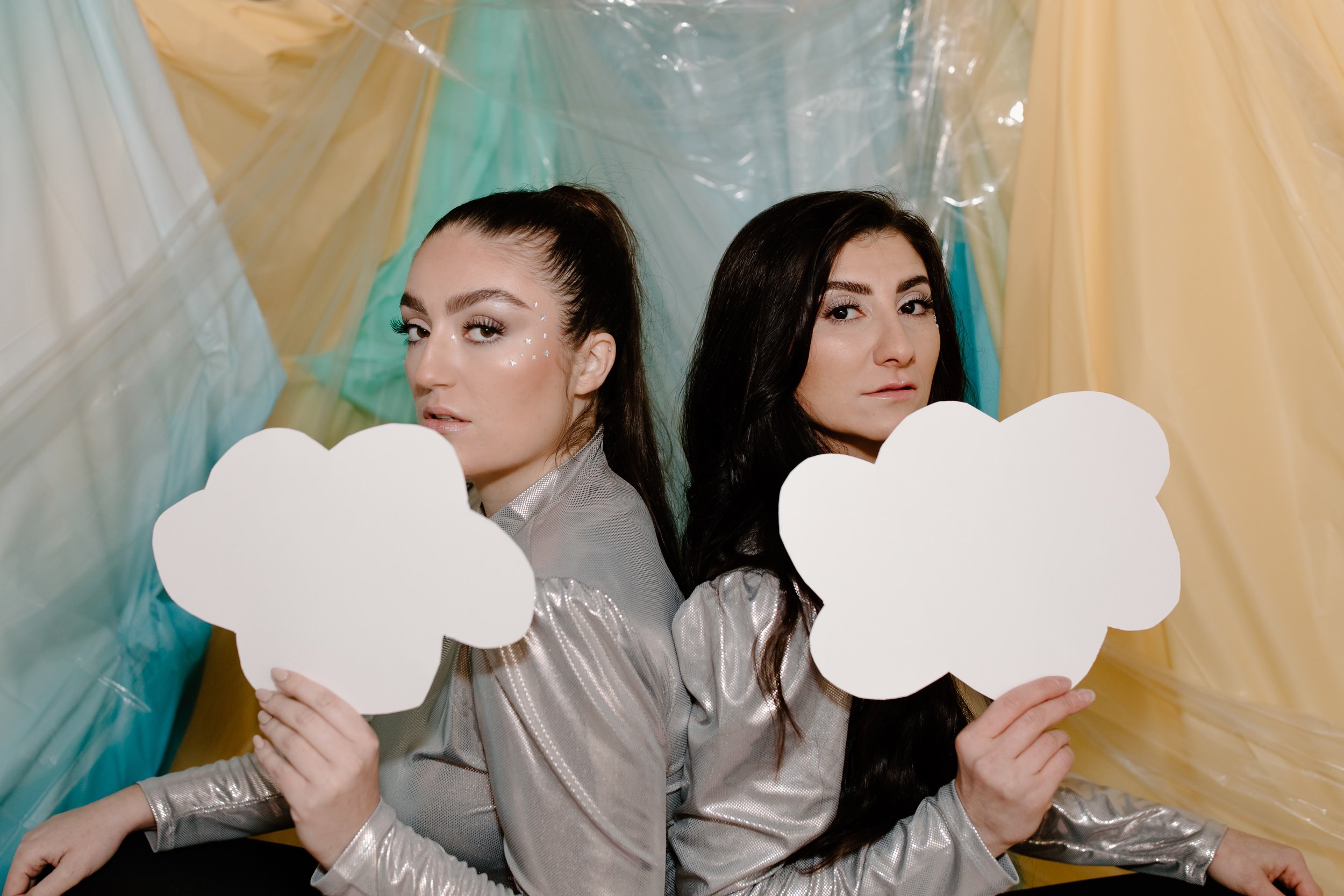 Two women with metallic silver jackets sitting back to back, each holding a white cloud-shaped cutout, with colorful fabric background.