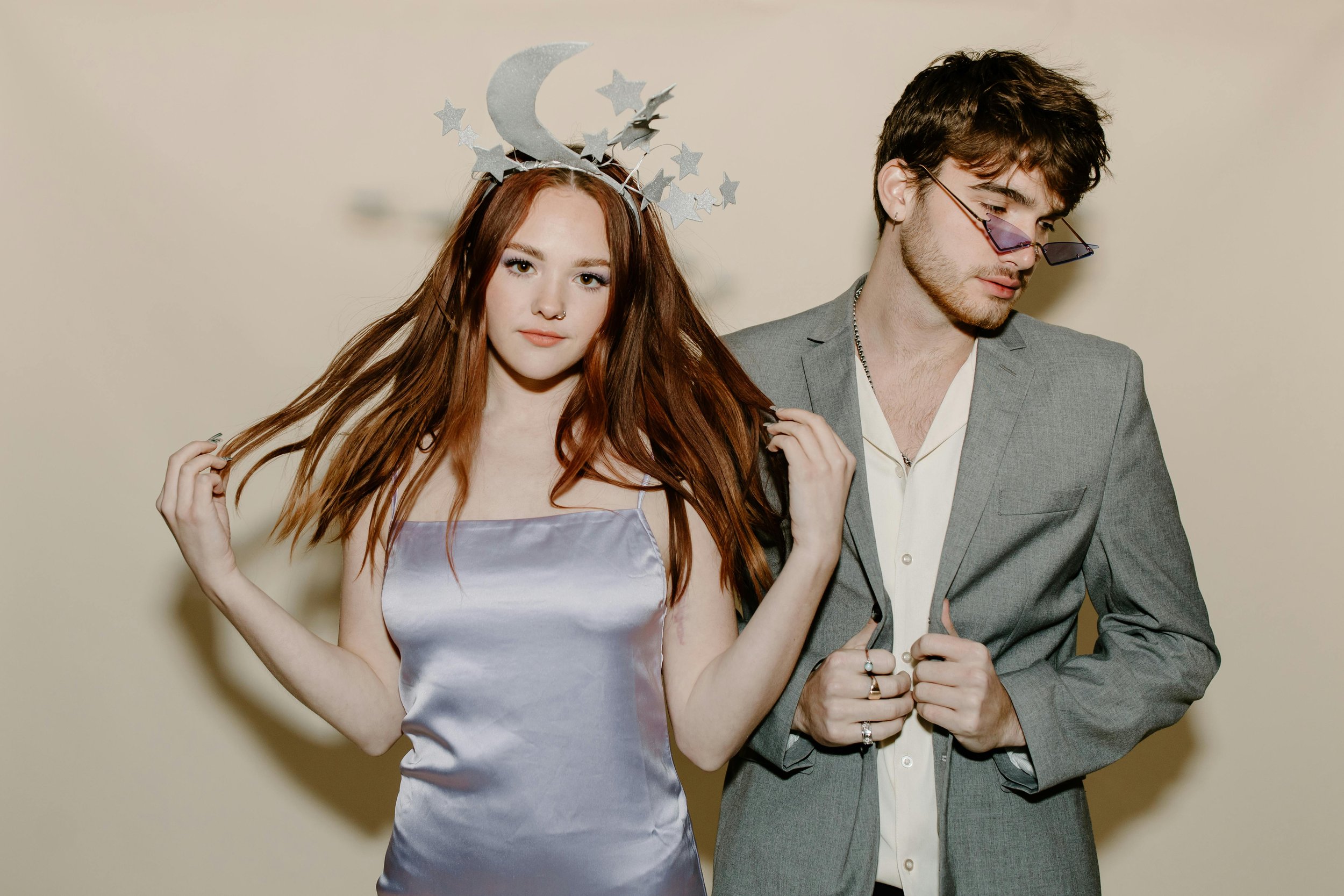 A young woman with long red hair and a young man with brown hair and glasses pose against a light beige background. The woman wears a silver satin dress and a moon and stars headband, while the man is dressed in a gray blazer with rings and necklaces.