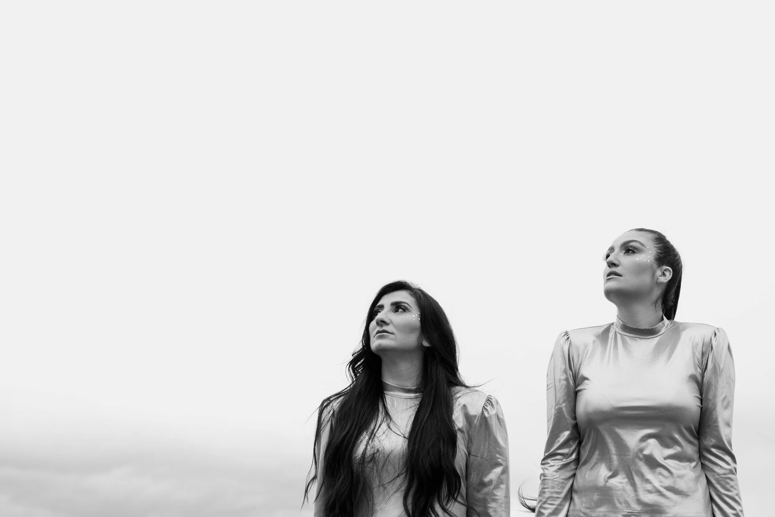 Two women with long hair and shiny dresses standing outdoors against a cloudy sky, looking upwards.