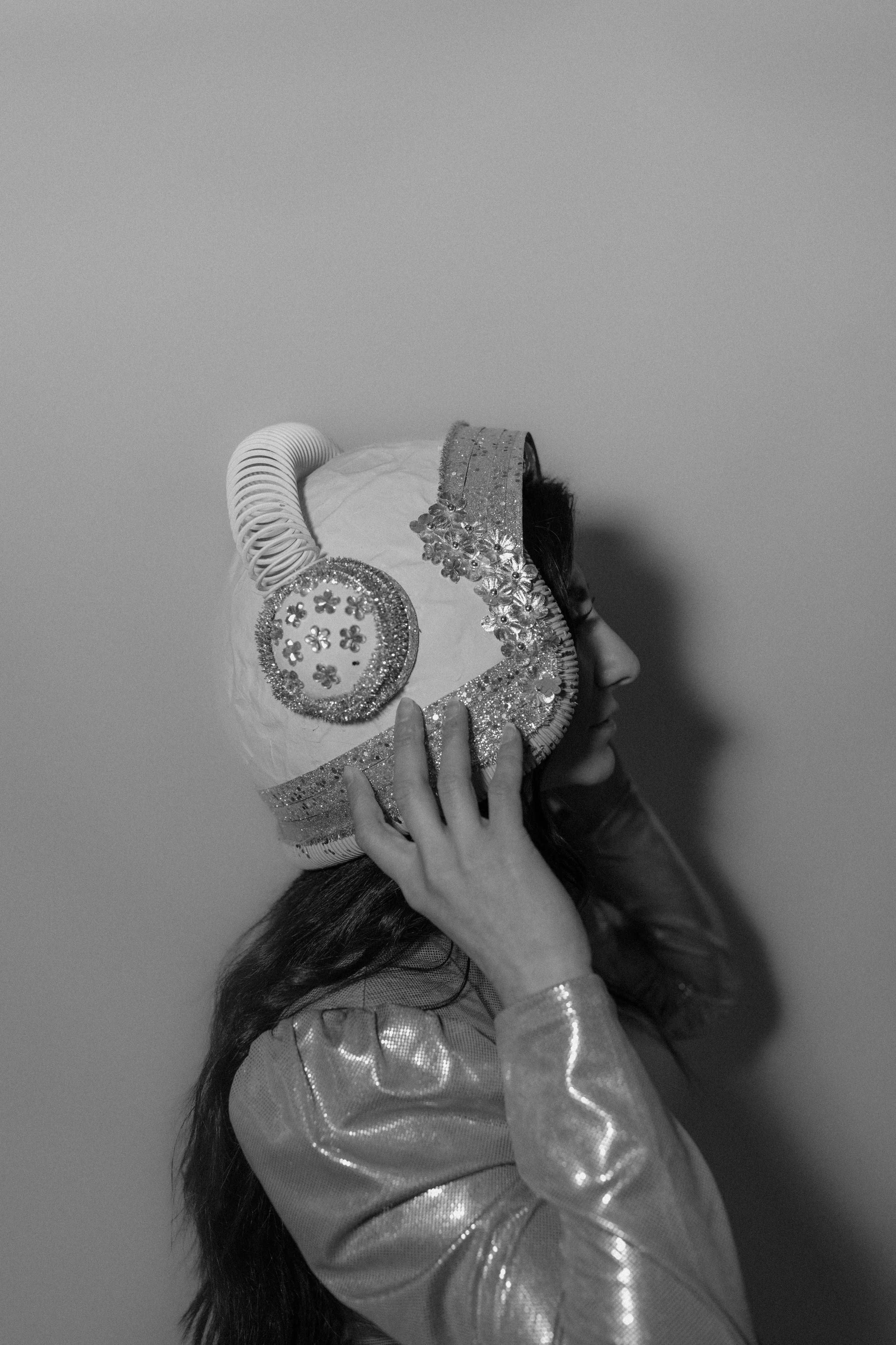 A woman wearing a shiny jacket and holding a decorated balloon in the shape of a head with horns and floral details, standing against a plain wall.