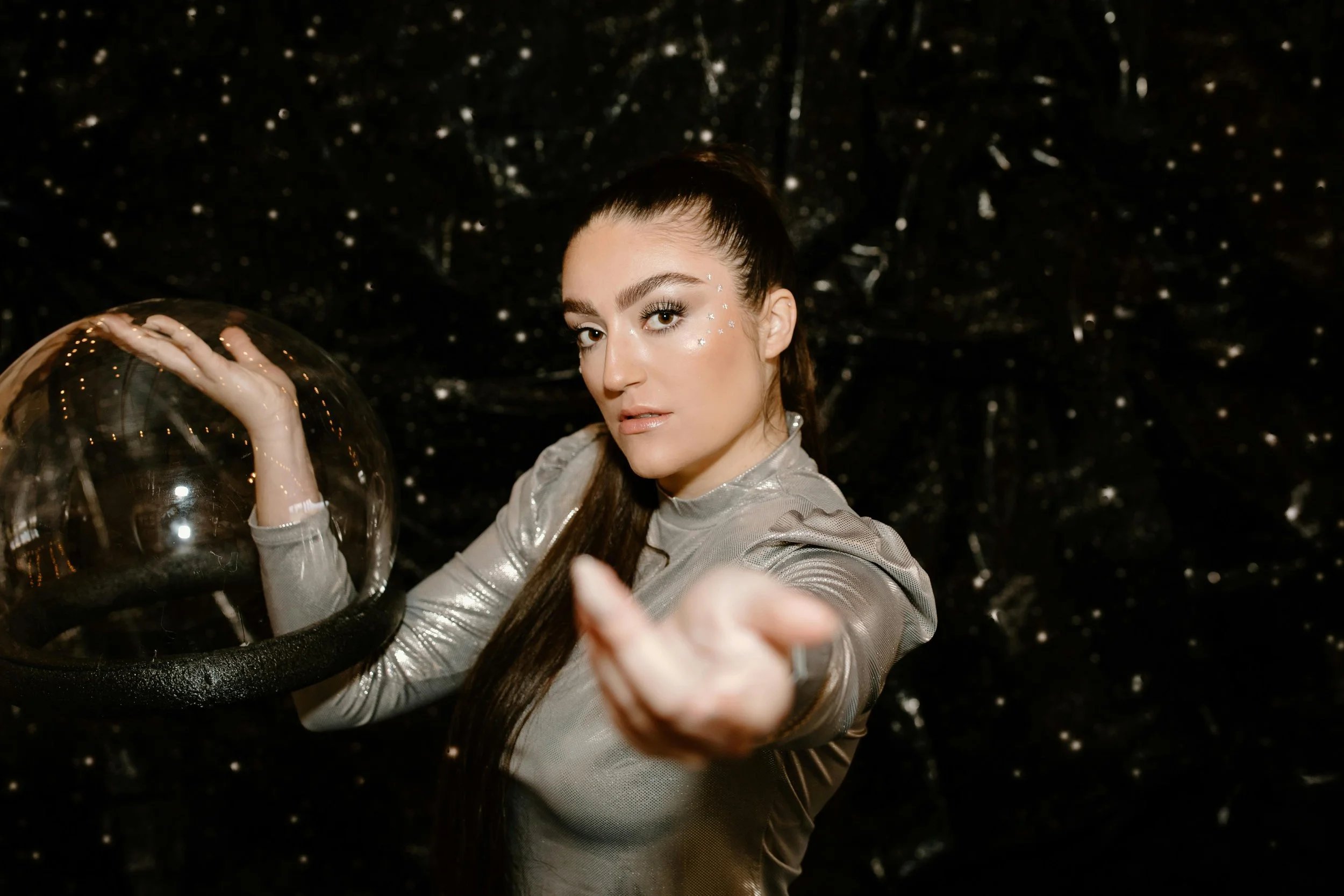 A woman in a shiny silver top posing with a transparent balloon in front of a black background with small sparkling lights.
