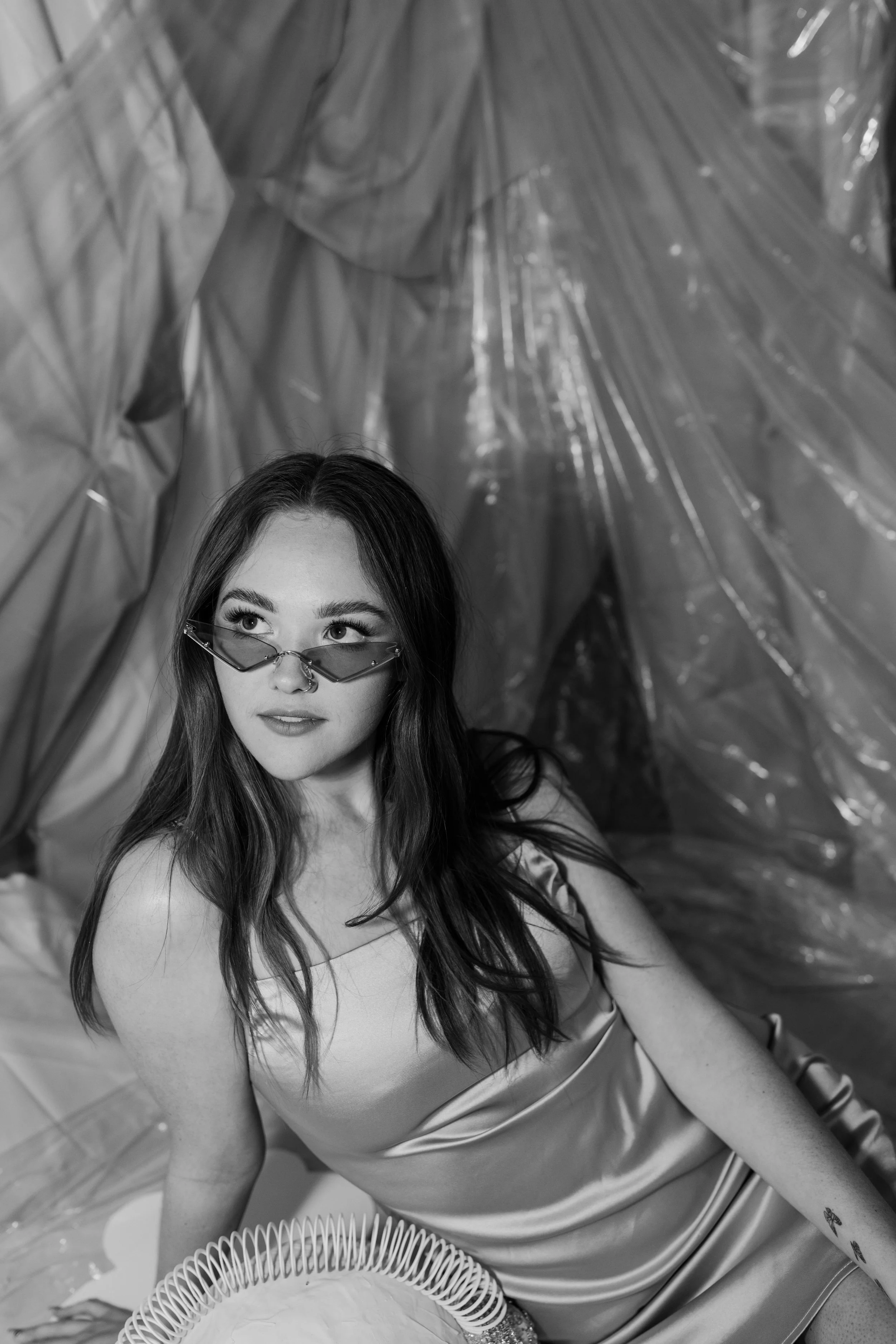 A young woman with long hair and sunglasses, wearing a satin dress, sitting in front of a backdrop made of plastic or fabric material.