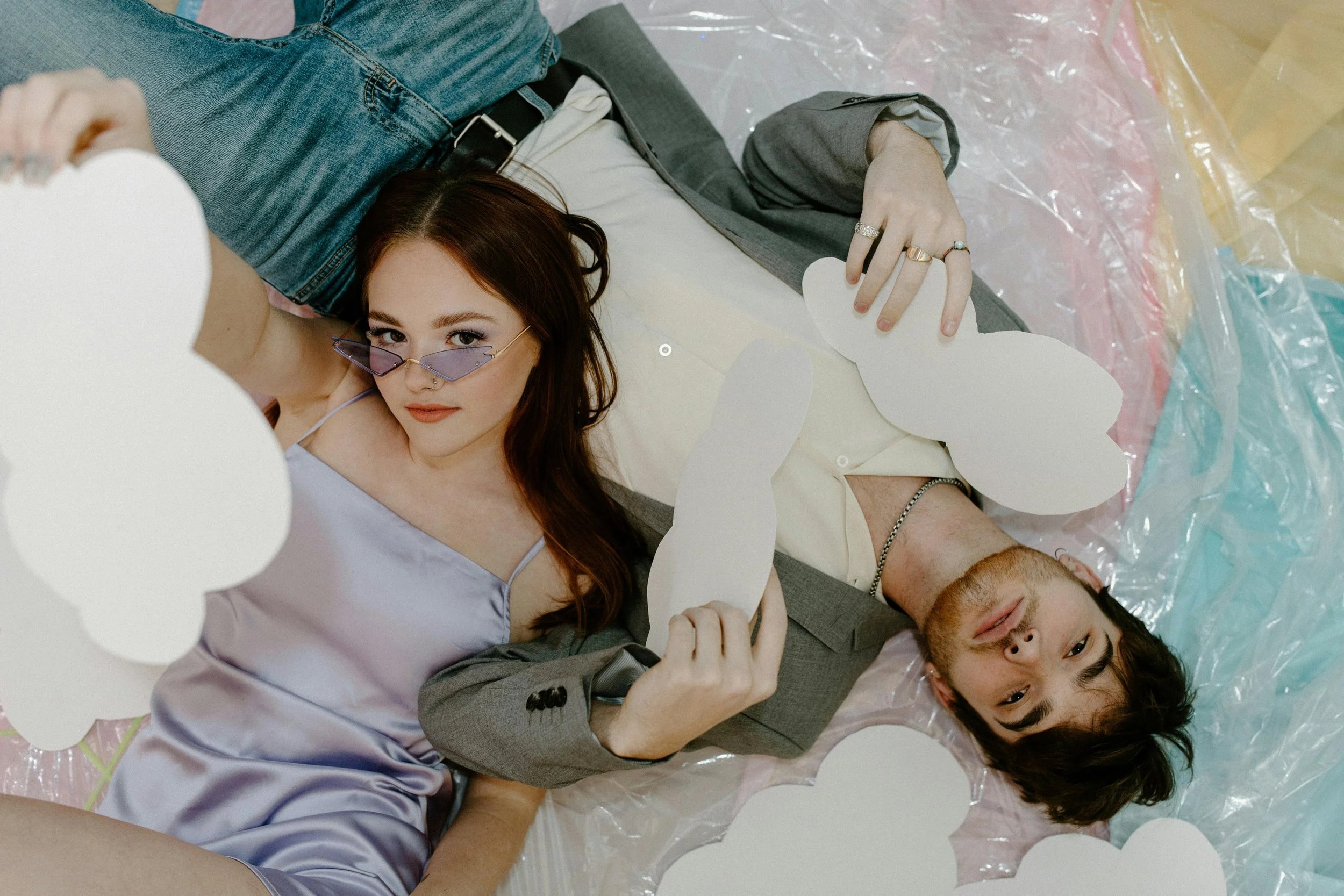 A woman with red hair and purple sunglasses lying on a colorful plastic surface next to a man with dark hair, both surrounded by white cloud-shaped cutouts.