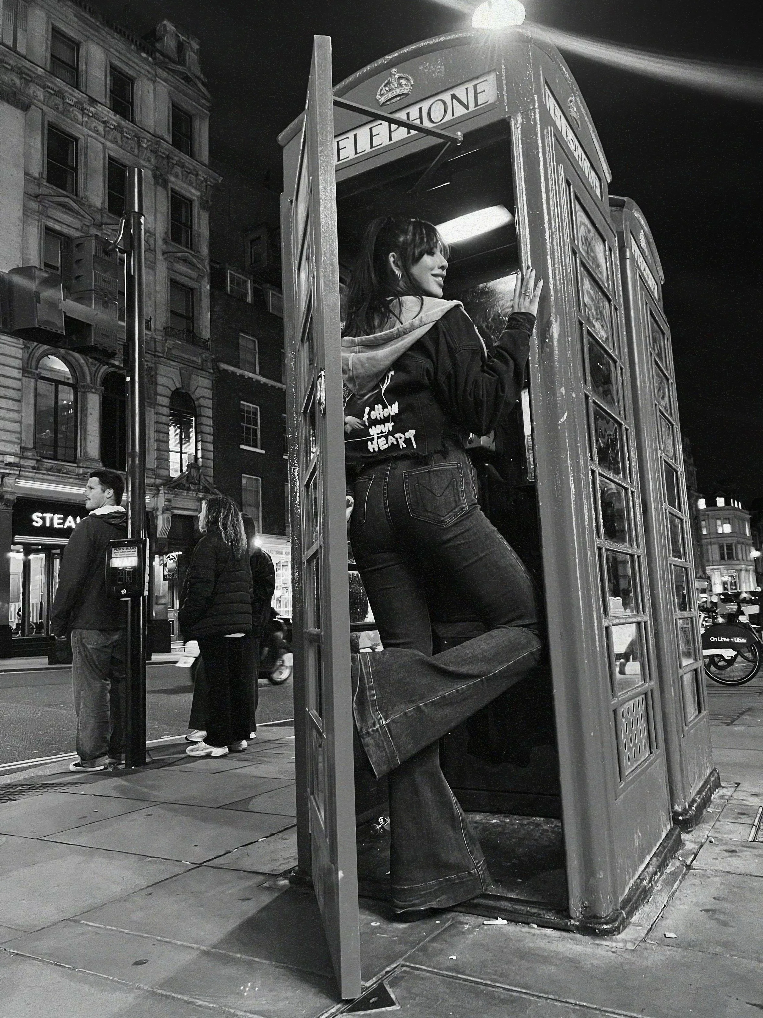 A young woman standing inside a classic British red telephone booth on a city street at night, with buildings and people in the background.