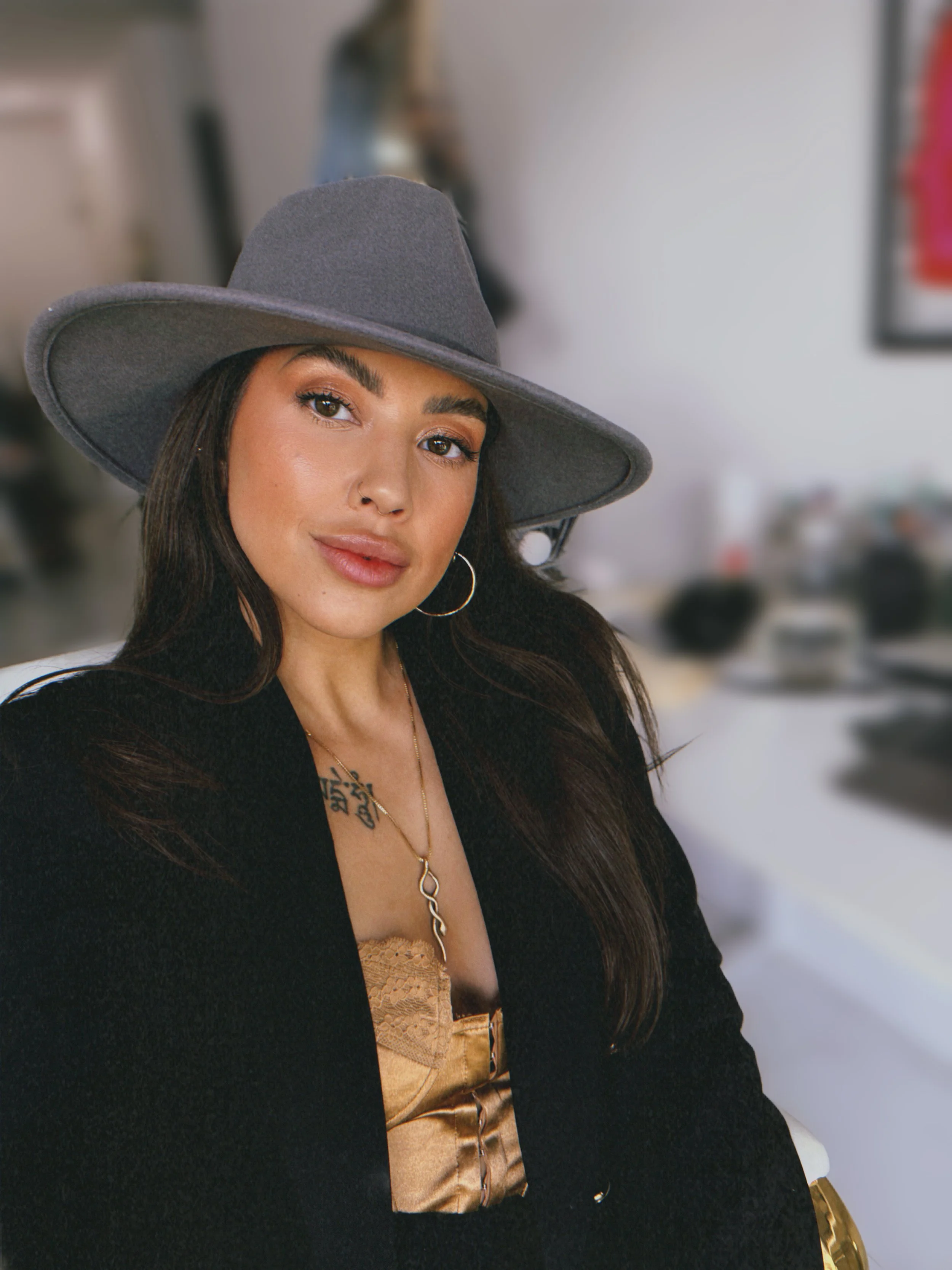 A woman with long dark hair, wearing a wide-brimmed gray hat, hoop earrings, a metallic gold top, and a black jacket, sitting indoors with a blurred background.