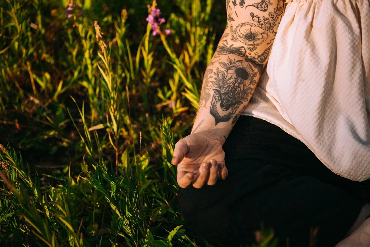 Jessica Huss seated in grass, tattooed arm resting on leg with palm up