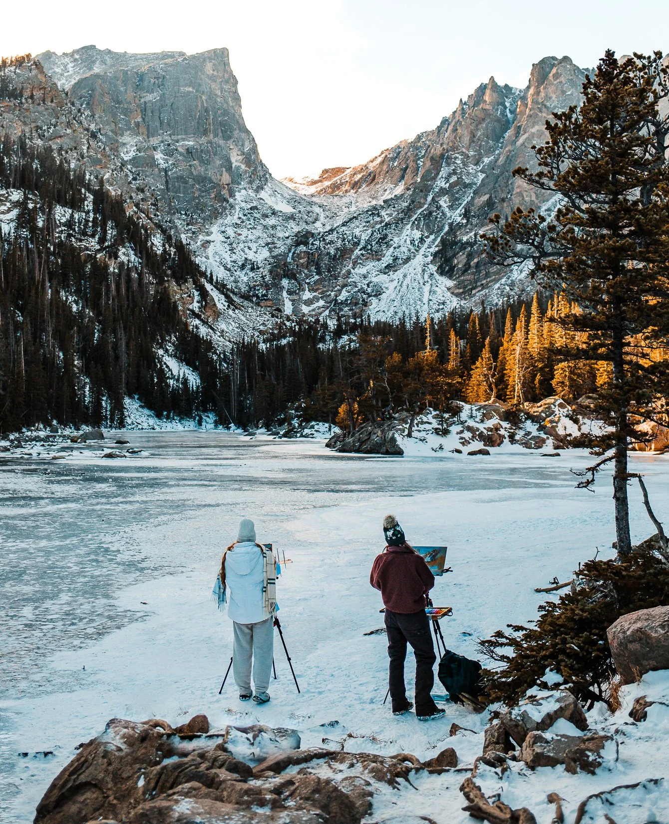 Two people in winter clothing are standing on snow-covered ground near a frozen lake, with mountains and snow-dusted trees in the background.