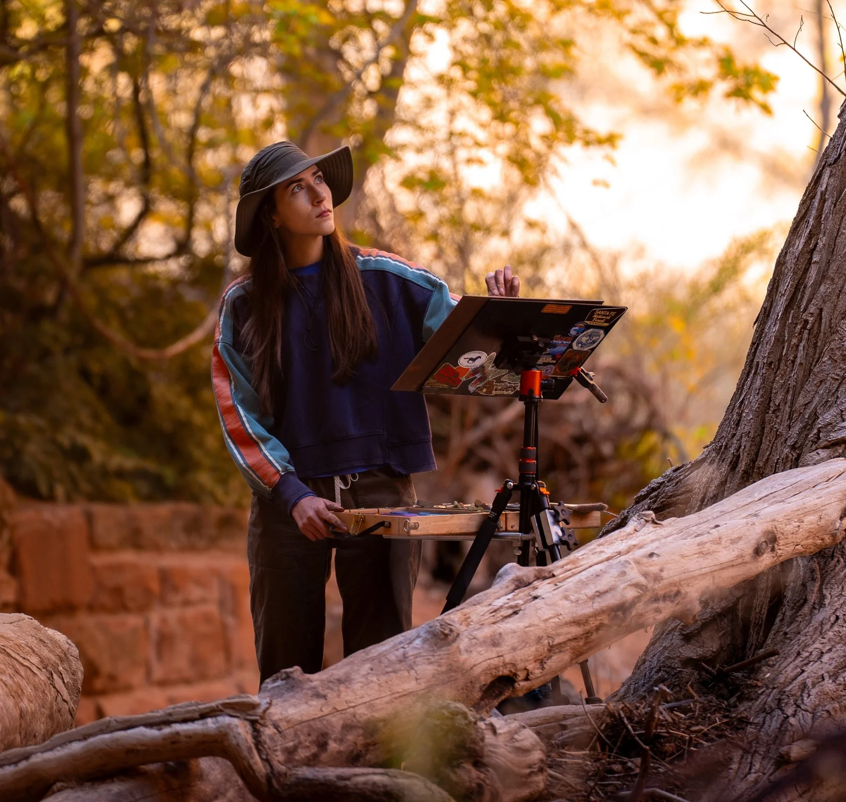 Julia Robinson, the plein artist, stands outdoors in a forested area during autumn, looking thoughtfully ahead as she paints in front of her plein air easel.