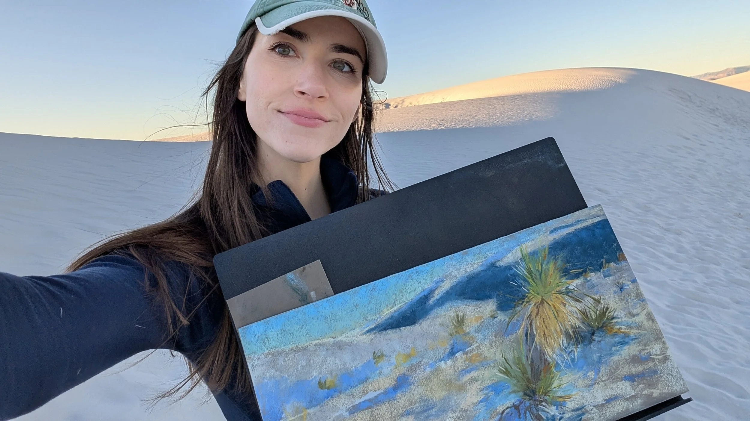 A woman with long brown hair, wearing a baseball cap, taking a selfie in a snow-covered desert landscape, holding paintings of a beach scene with palm trees.