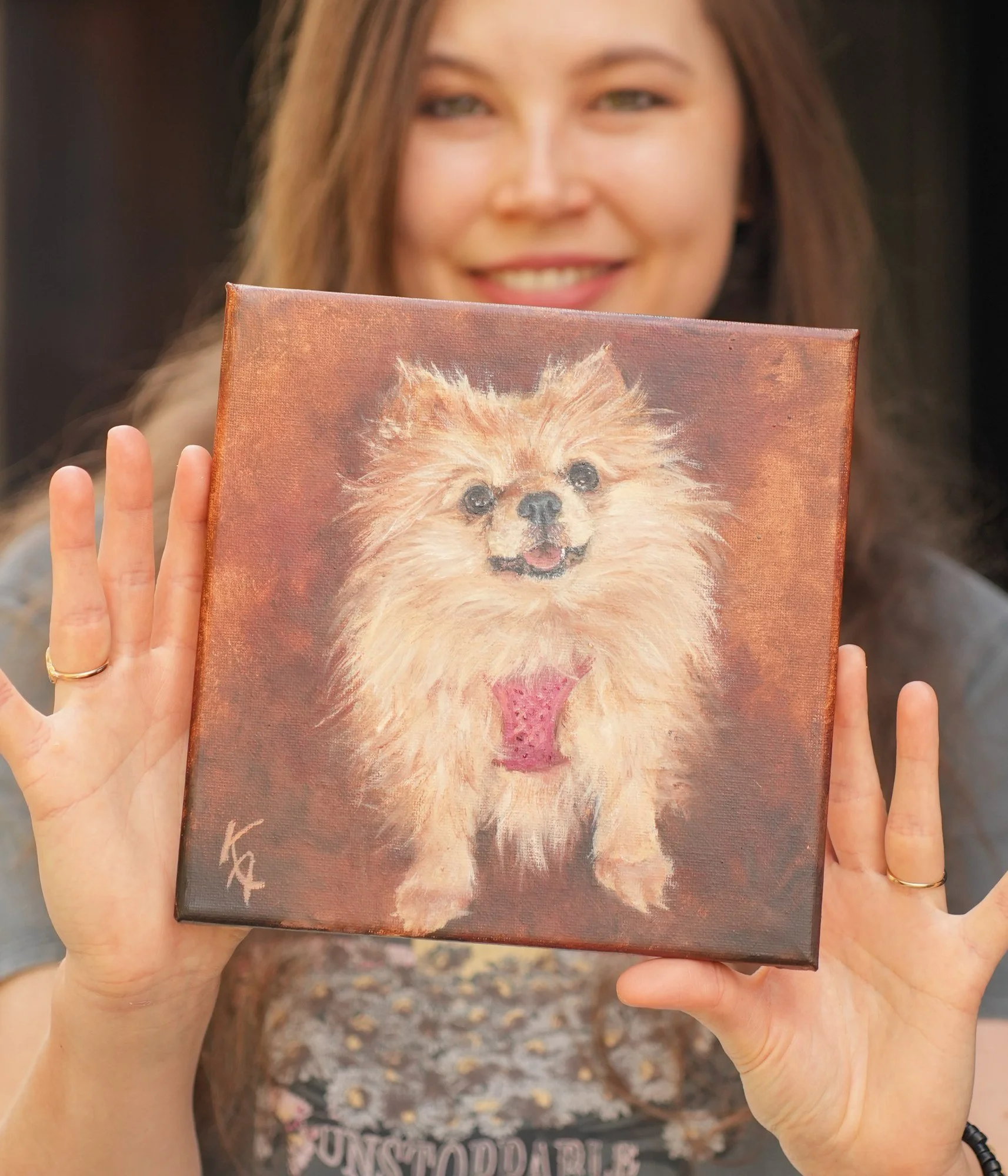 Artist smiling and holding a small oil painting of a Pomeranian dog with pink bow, warm brown background