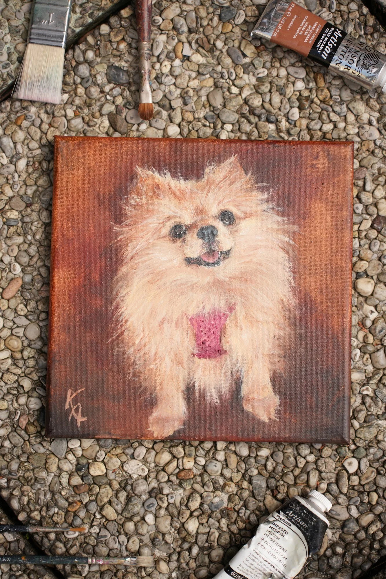 Pomeranian art oil painting on gravel surrounded by paintbrushes and paint tubes