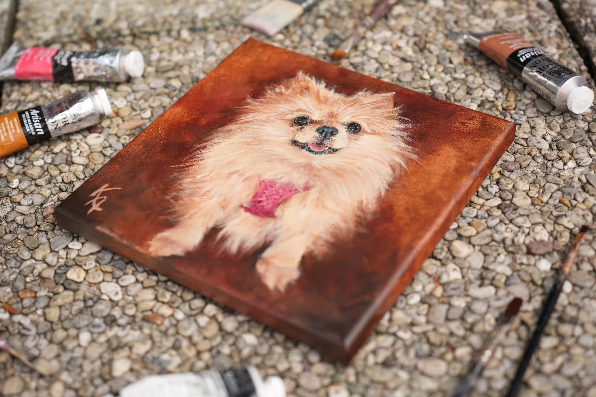 Oil painting of a fluffy Pomeranian dog with pink bow, photographed outdoors on gravel next to paint tubes and brushes