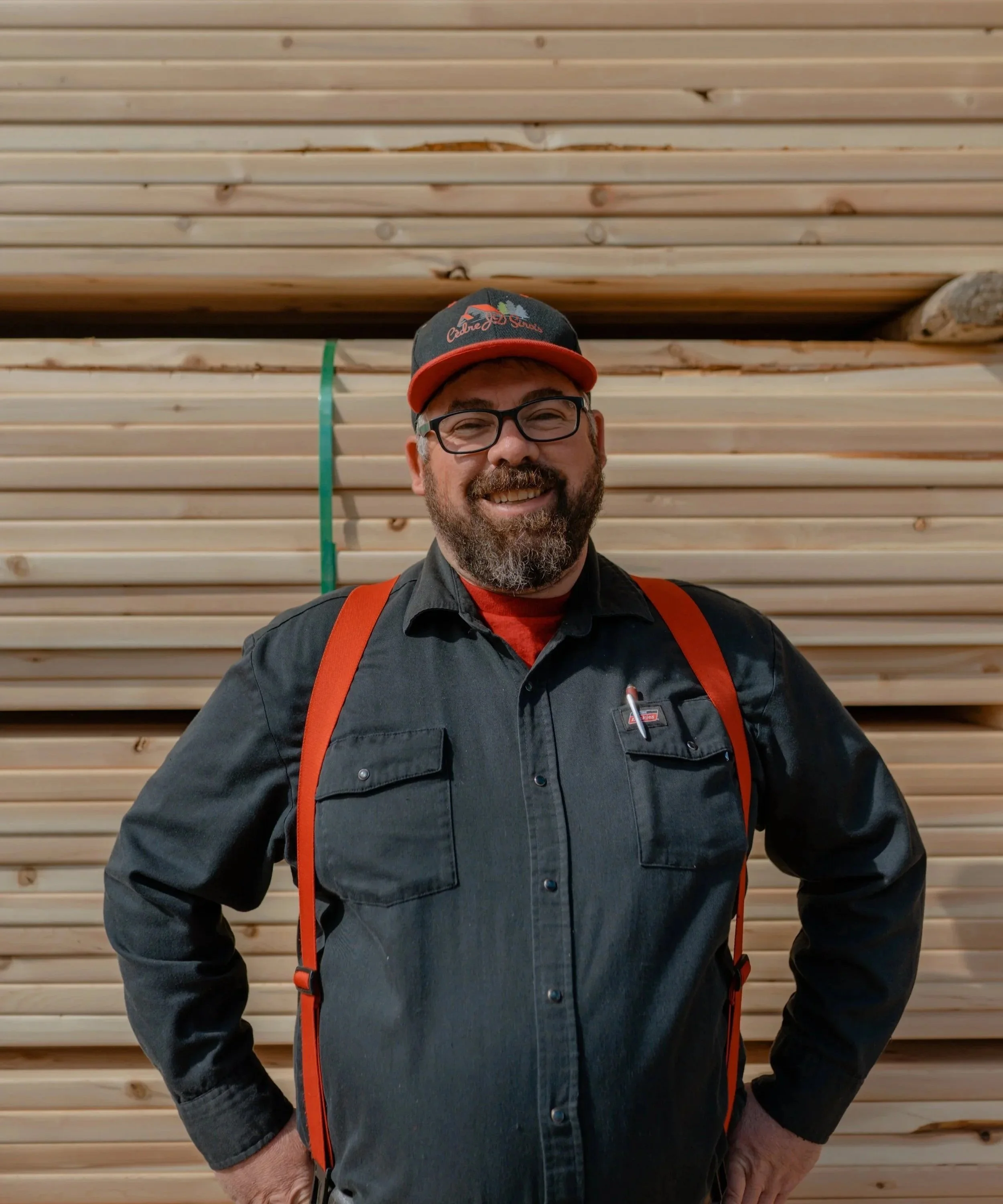 Un homme souriant portant une casquette, des lunettes, une chemise noire et un sac à dos orange, debout devant un mur de planches en bois.