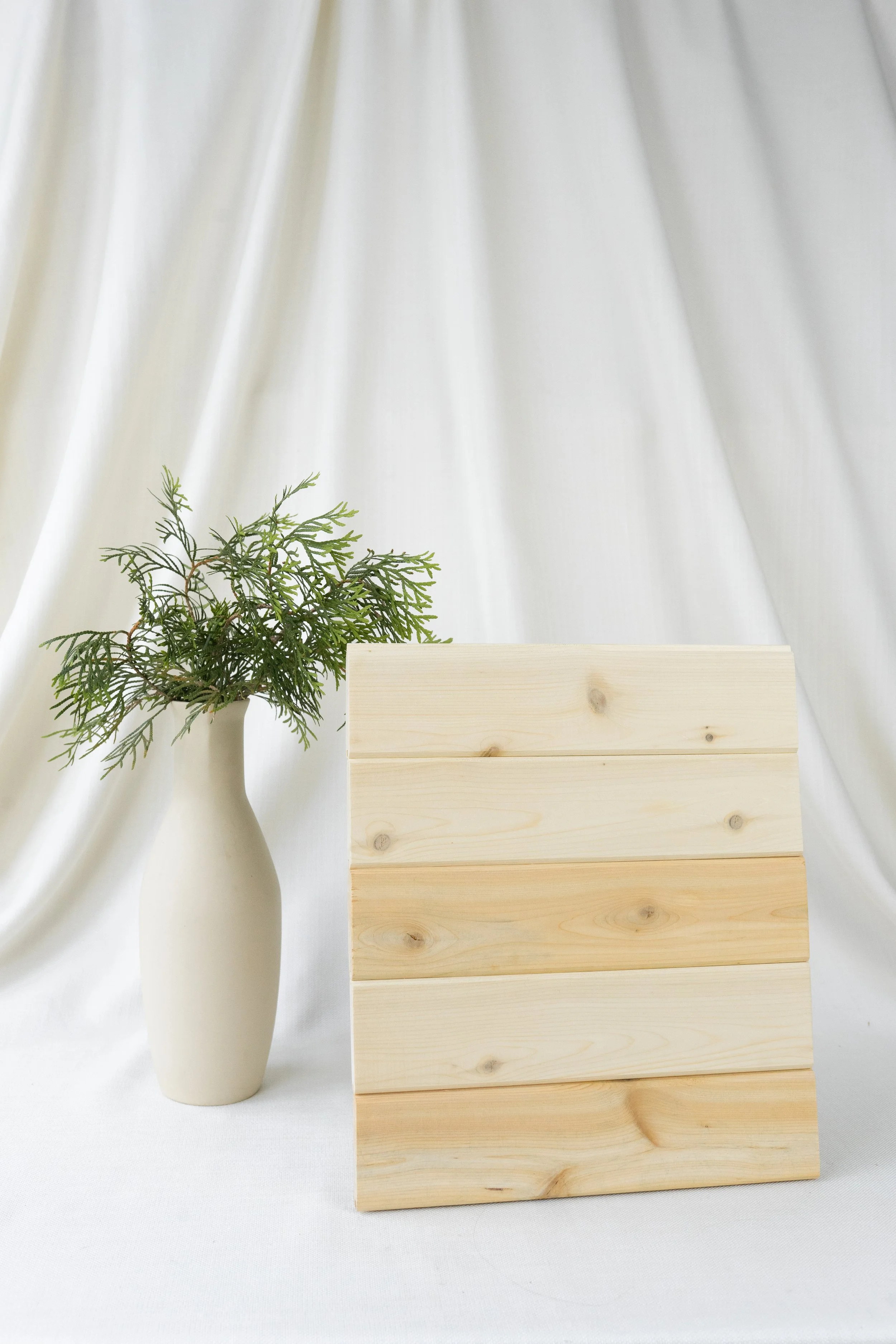 Vase blanc avec des branches de feuillage vert à côté d'un panneau en bois clair contre un fond de rideau blanc.