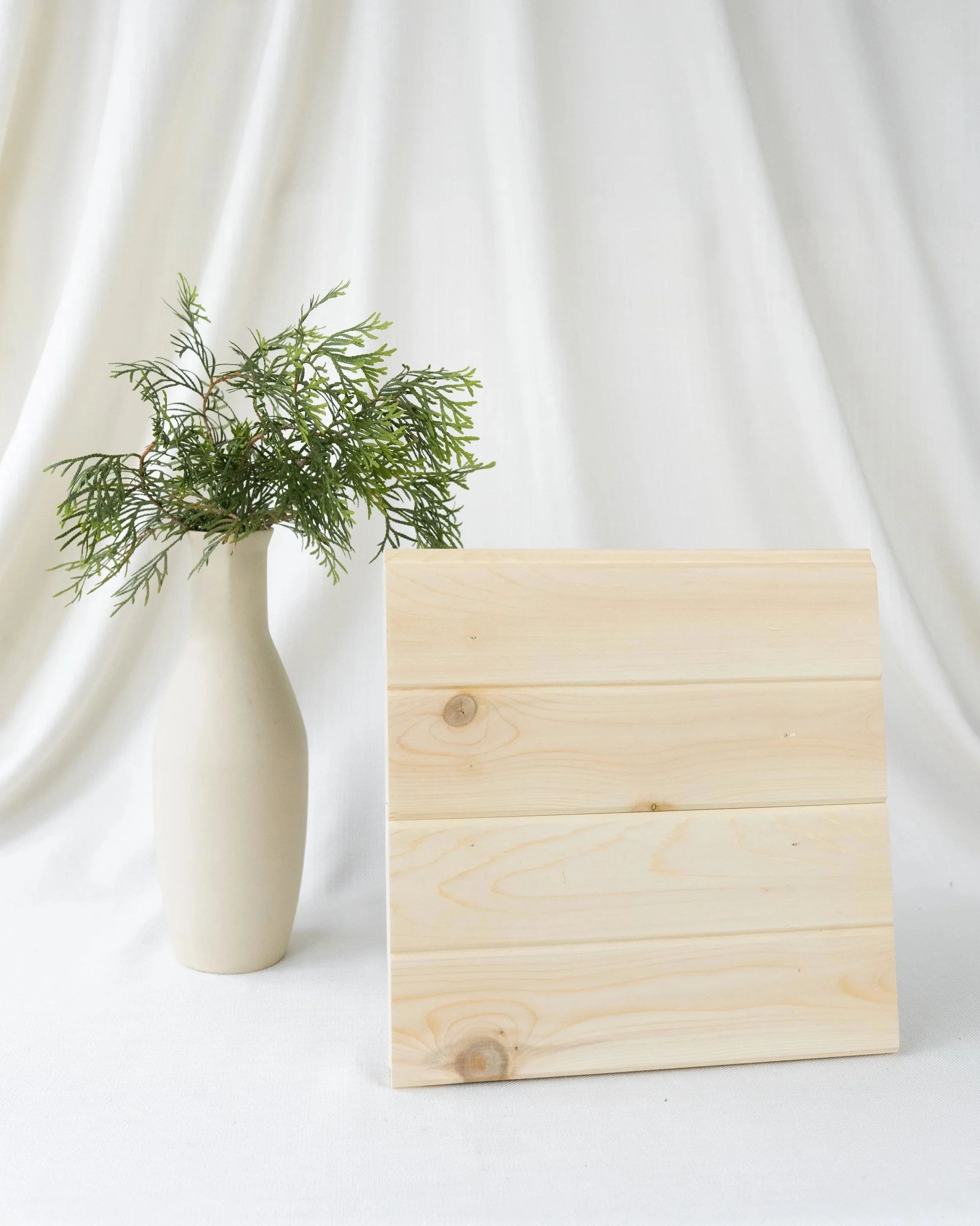 Vase blanc avec des branches de feuillage vert à côté d'un panneau en bois clair contre un fond de rideau blanc.