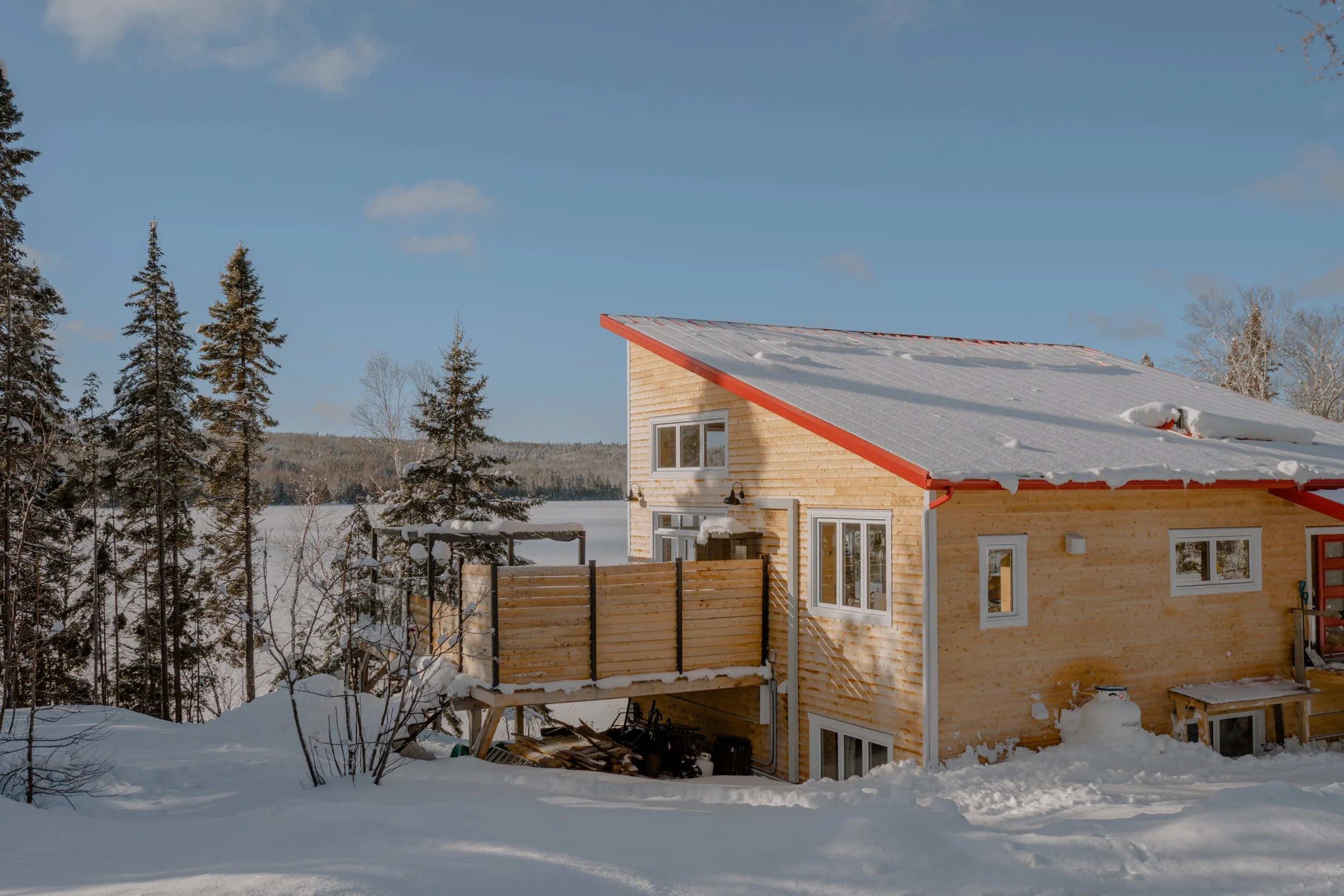 Maison en bois avec une terrasse en bois, entourée de neige, dans un paysage enneigé avec des arbres et un ciel bleu.