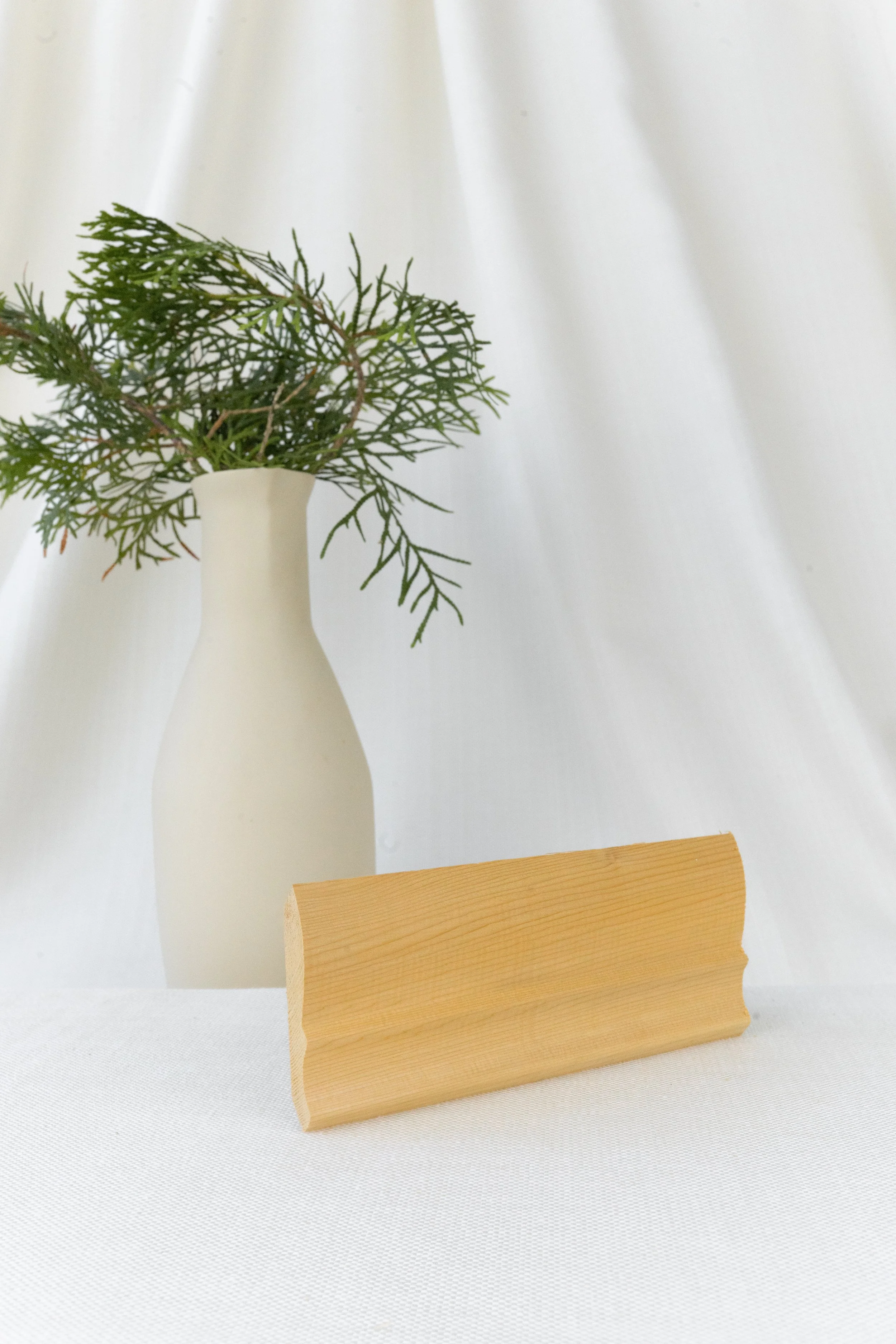 une vase blanche contenant des branches vertes, un morceau de bois sur une table en tissu blanc, avec un fond blanc