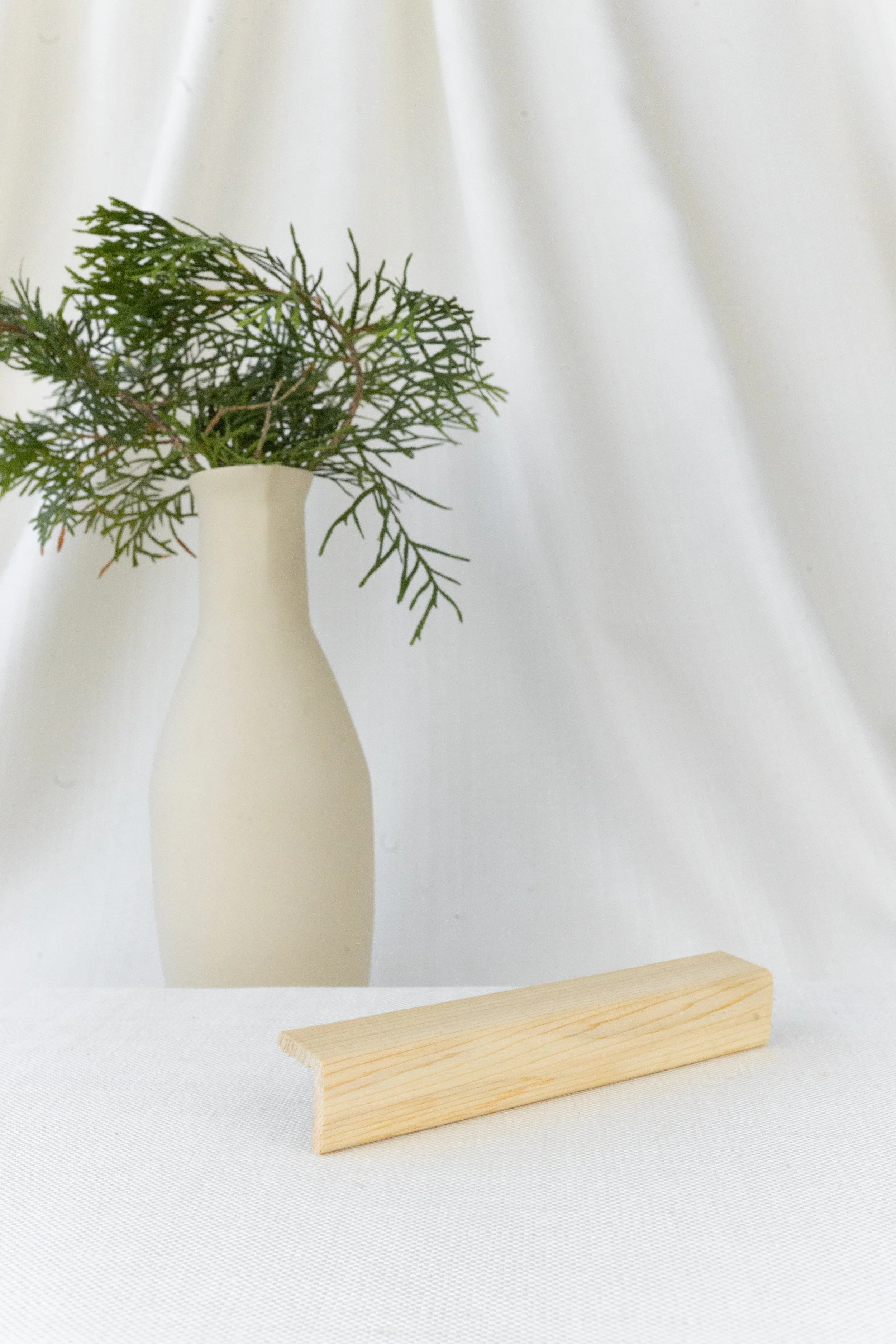 Une petite branche verte dans un vase blanc en arrière-plan, avec une planche de bois sur une surface blanche, fond blanc avec tissu.