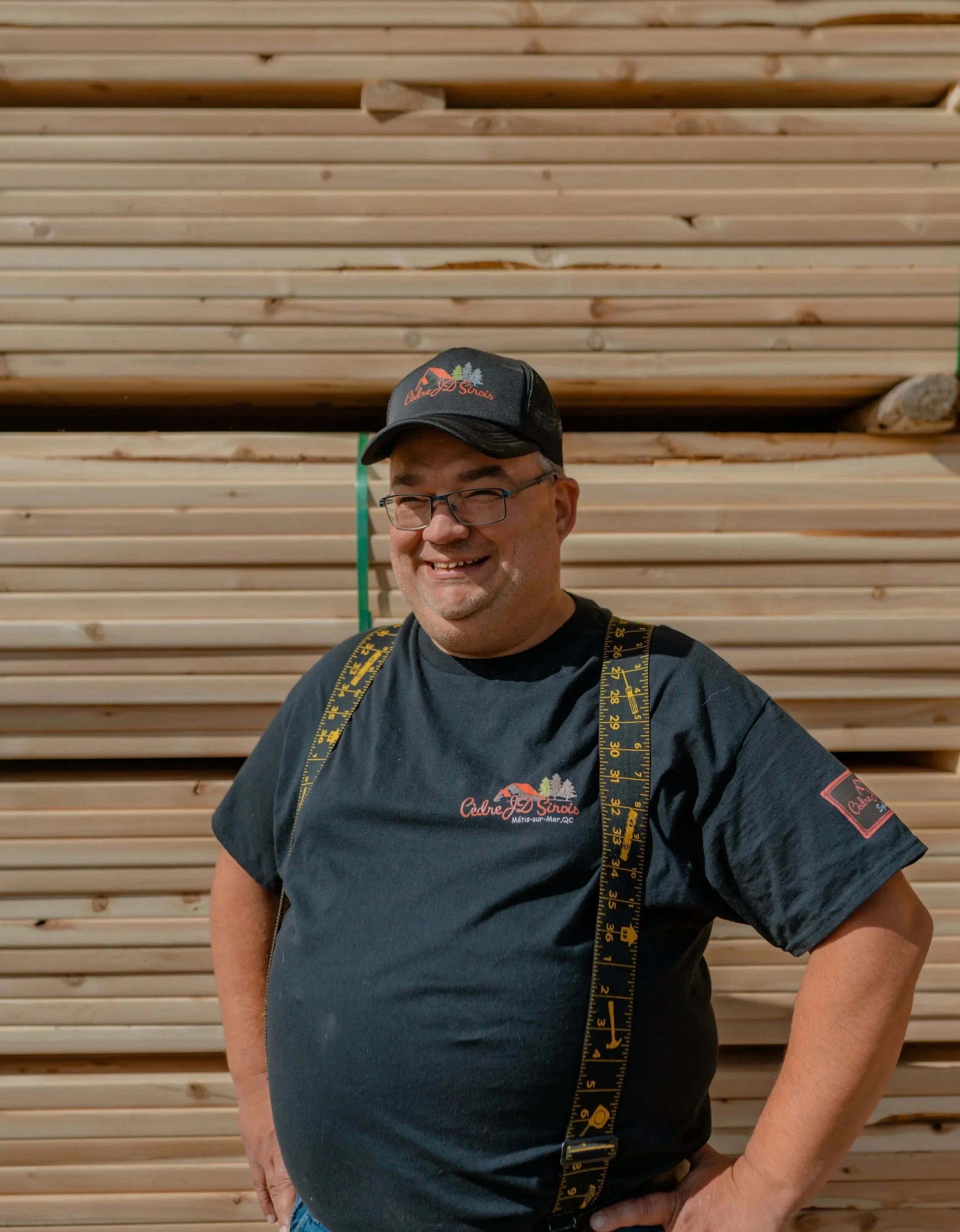 Un homme souriant portant un t-shirt noir avec un logo, un chapeau noir avec un logo, des lunettes, et une ceinture à mesurer enroulée autour du cou, avec un fond de planche en bois.