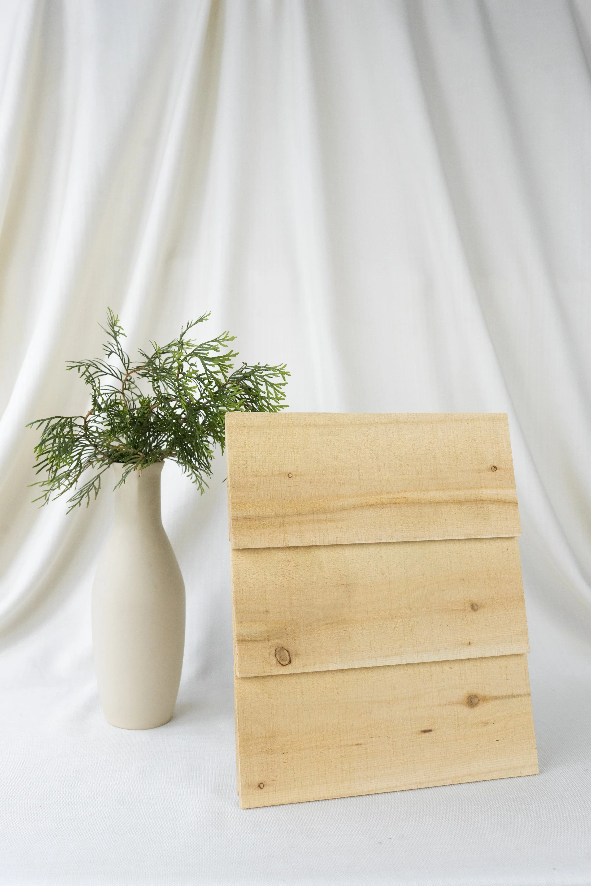 Une vase blanche avec une branche de feuillage vert à côté d'un panneau en bois naturel avec un fond de drap blanc.