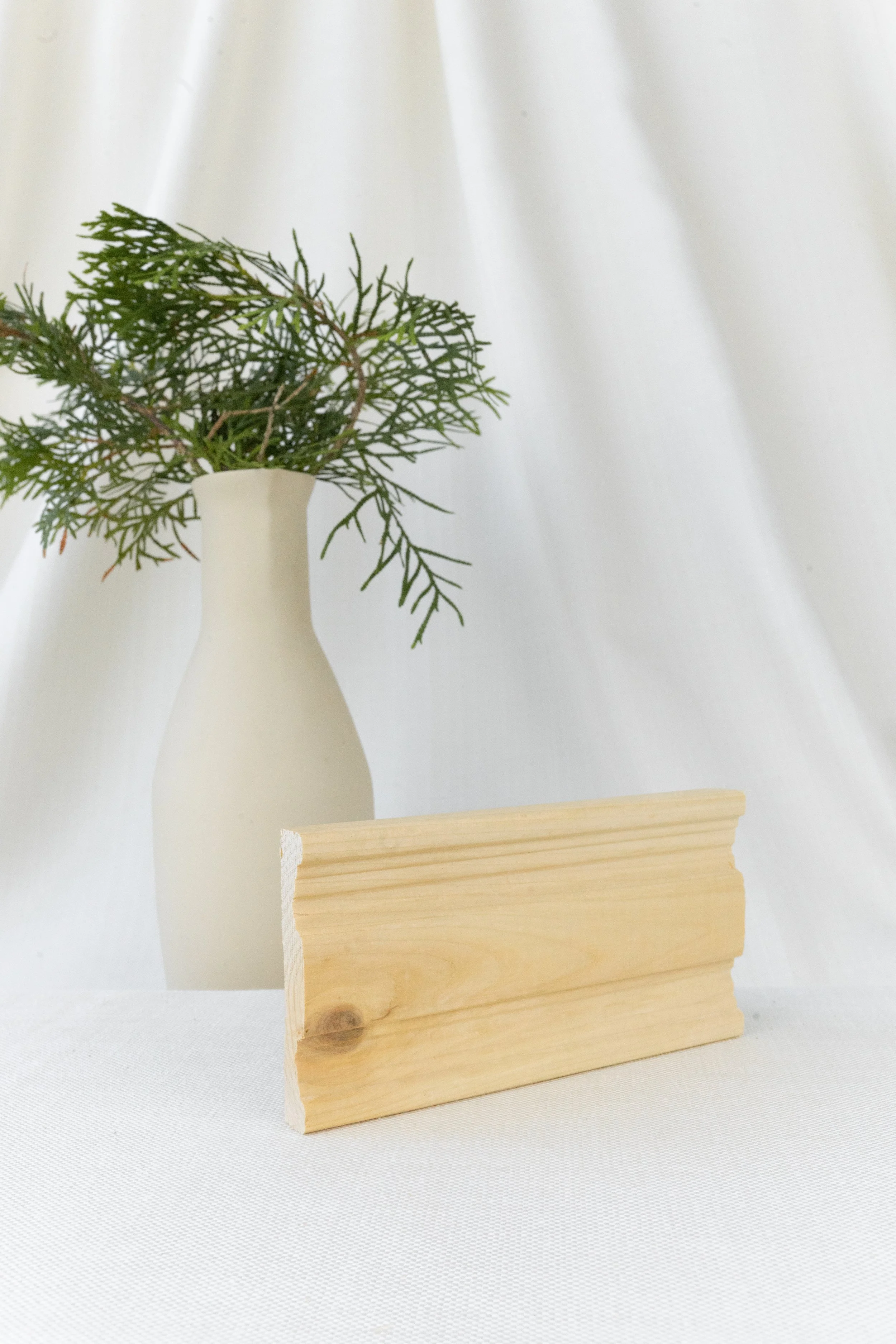 Une vase blanche avec des branches vertes, posée sur une surface blanche, en arrière-plan un rideau blanc.