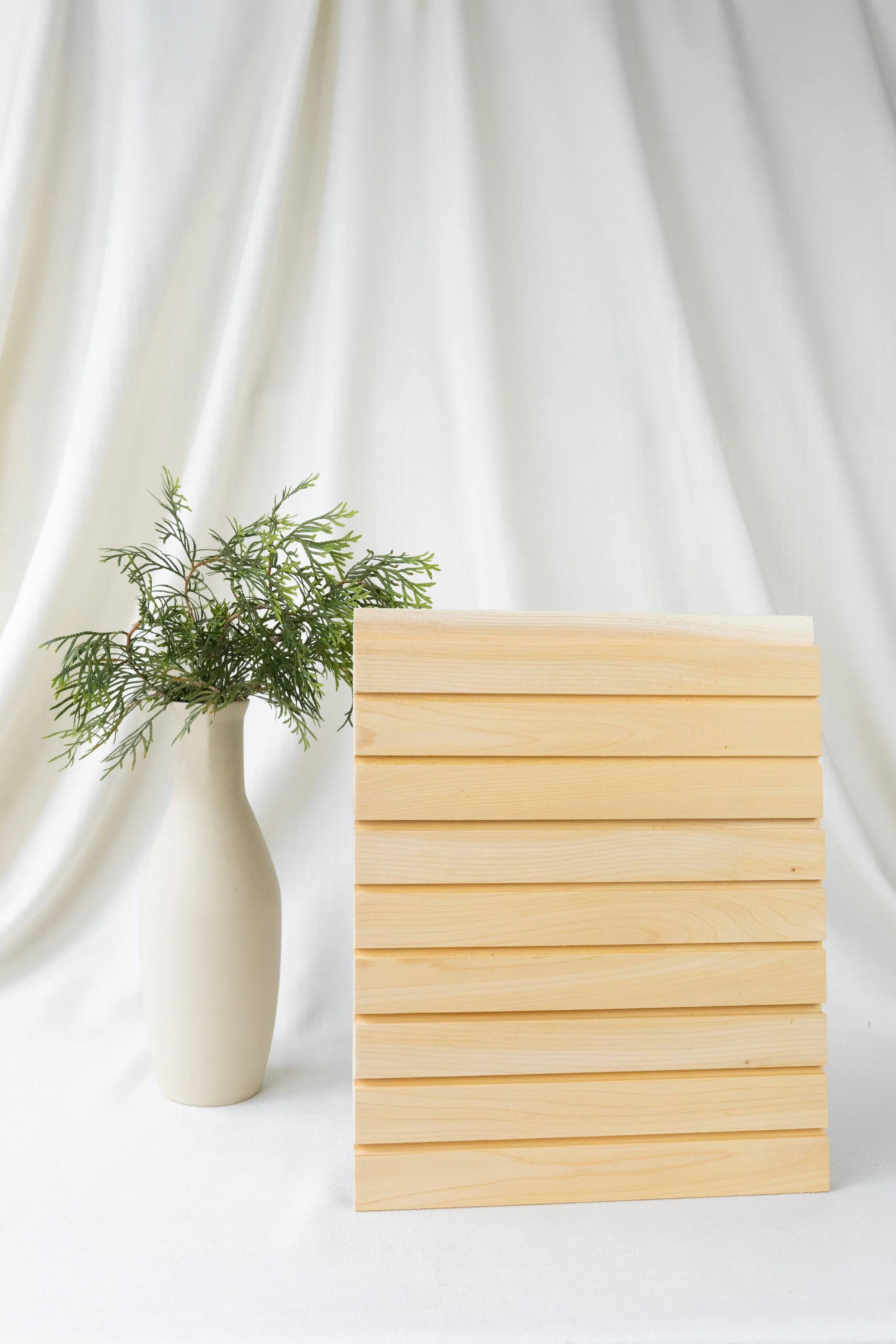 Une vase blanche avec une branche de feuillage vert à côté d'une pile de planches en bois clair, sur un fond de tissu blanc