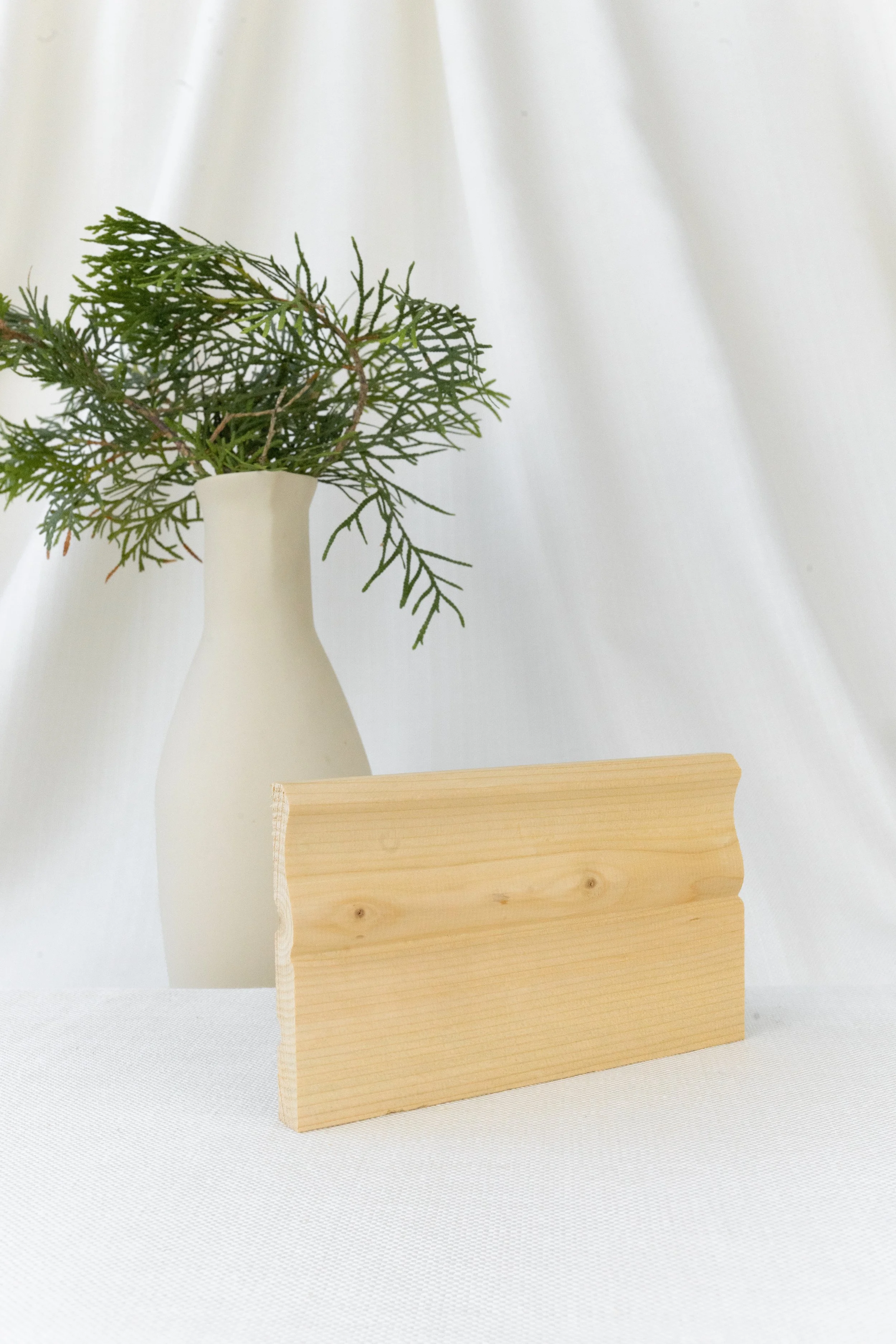 Vase blanc avec branches vertes en arrière-plan et planche de bois claire devant, fond de tissu blanc