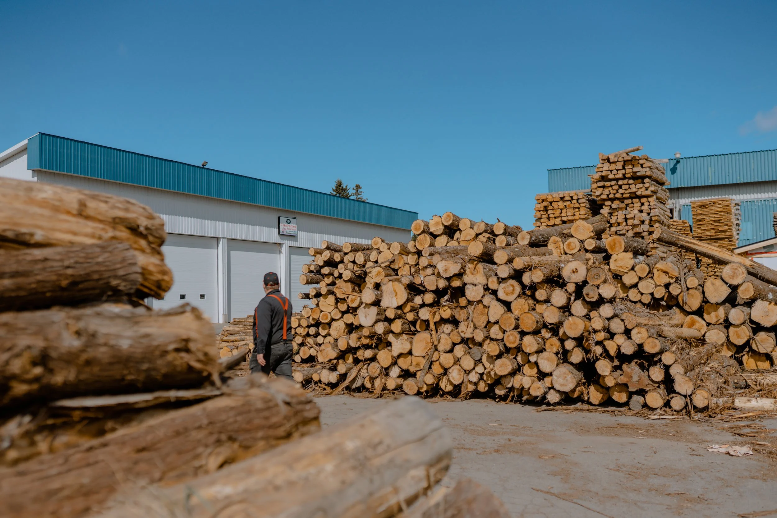 Un homme marche à côté d'une grande pile de bois coupé, devant un bâtiment industriel avec un toit bleu et des portes blanches, par une journée ensoleillée.