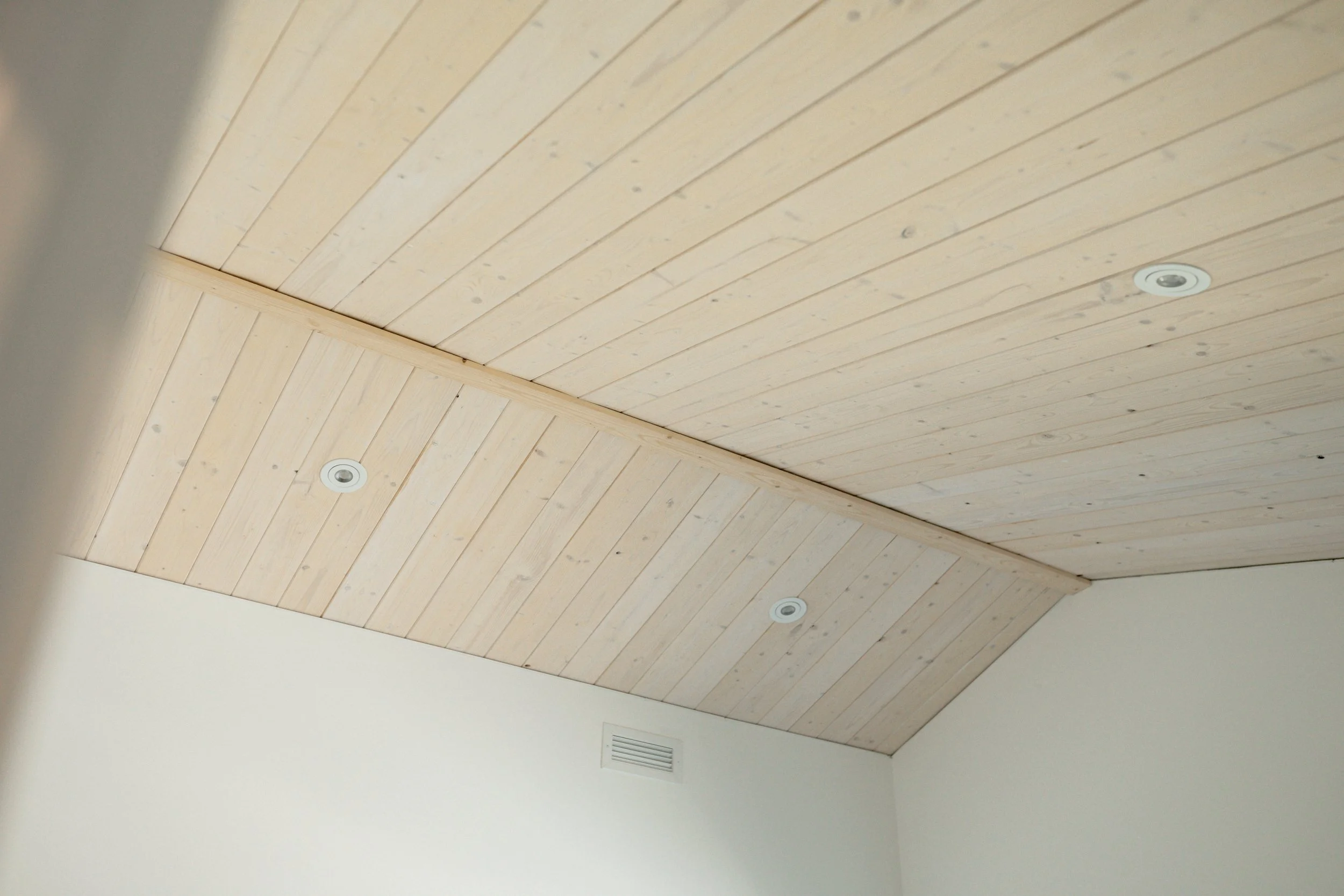 Plafond en bois clair avec trois spots encastrés, dans une pièce avec un mur blanc et une bouche d'aération.