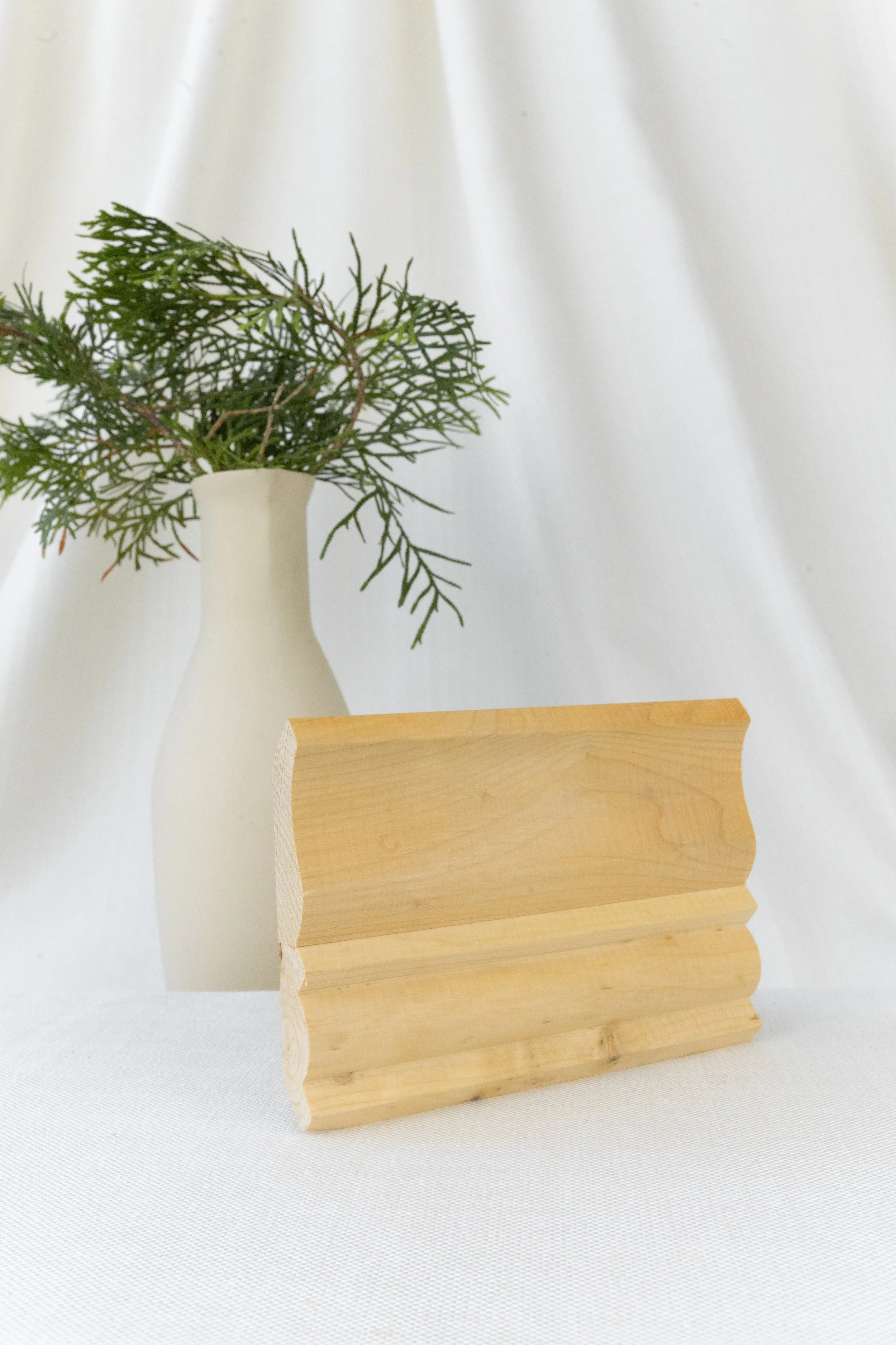 Vase blanche contenant des fougères, posé devant un fond blanc et un objet en bois