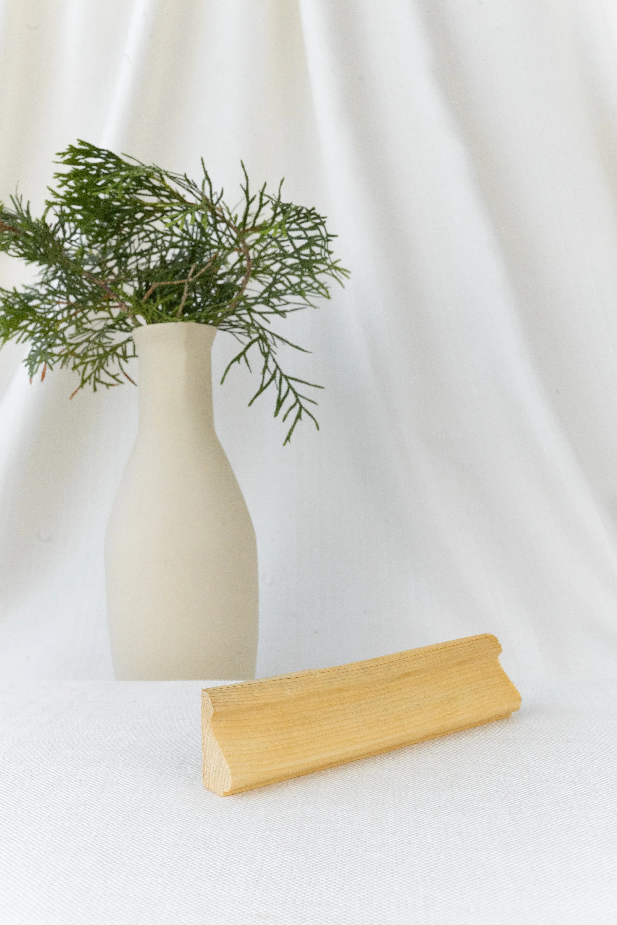 Une vase blanche avec des branches de feuillage vert, posée derrière un morceau de bois sur une surface blanche, avec un fond de tissu blanc.