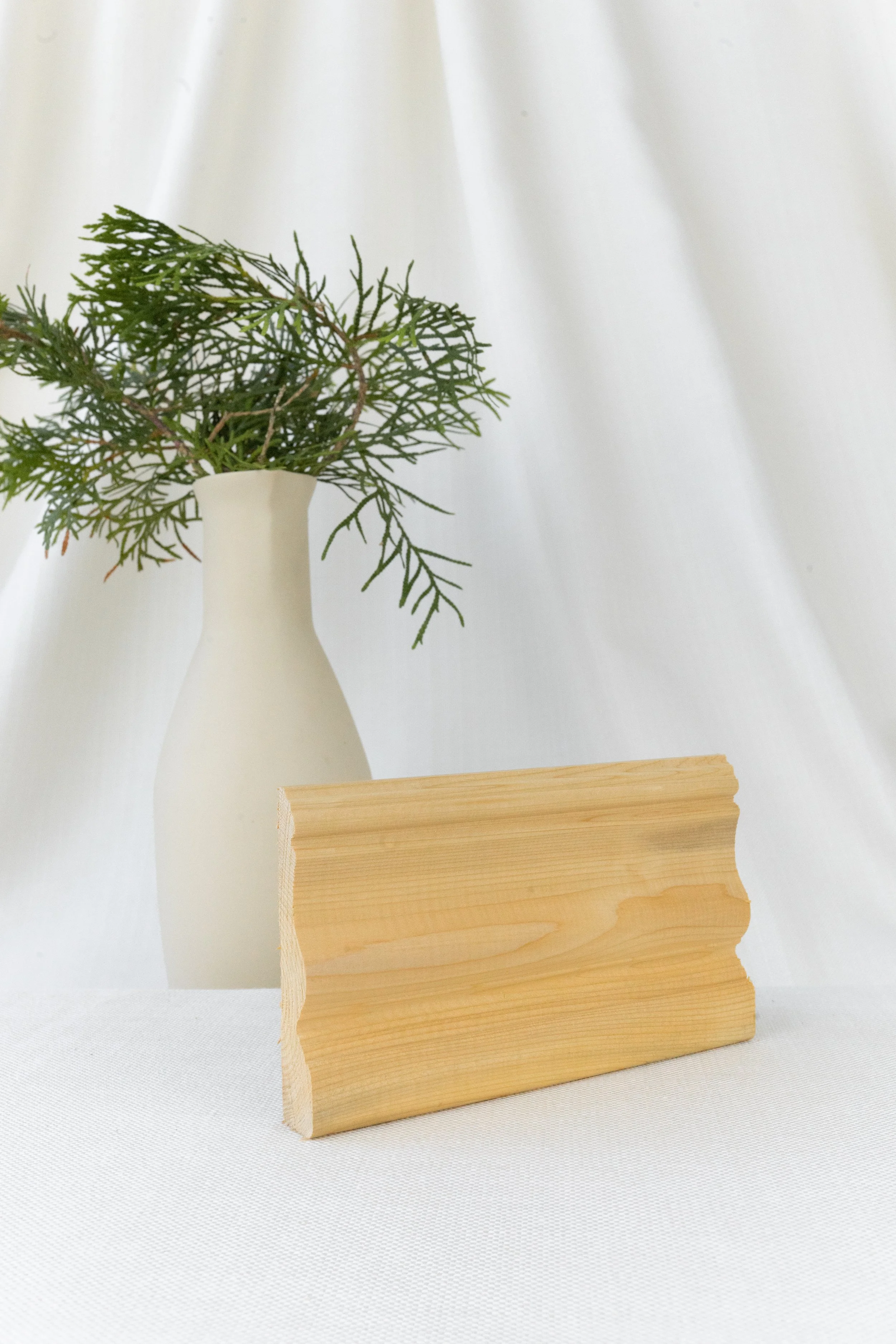 Vase blanc avec des branches vertes, panneau en bois clair, fond blanc et léger.