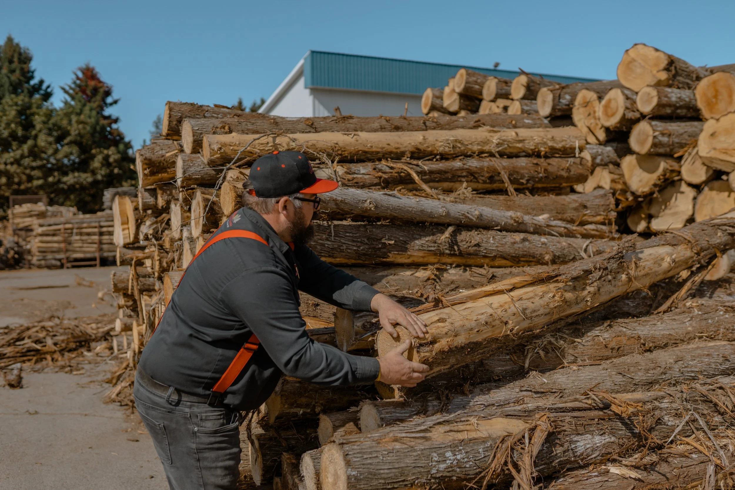 Un homme portant une casquette noire et une veste sombre coupe un morceau de bois dans une pile de bois empilé.